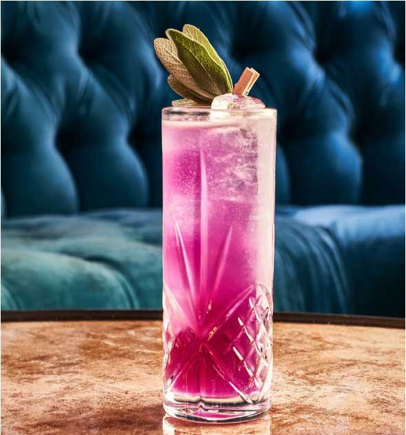 Vibrant pink cocktail in a tall glass with a sage garnish, on a reflective table, blue tufted background.