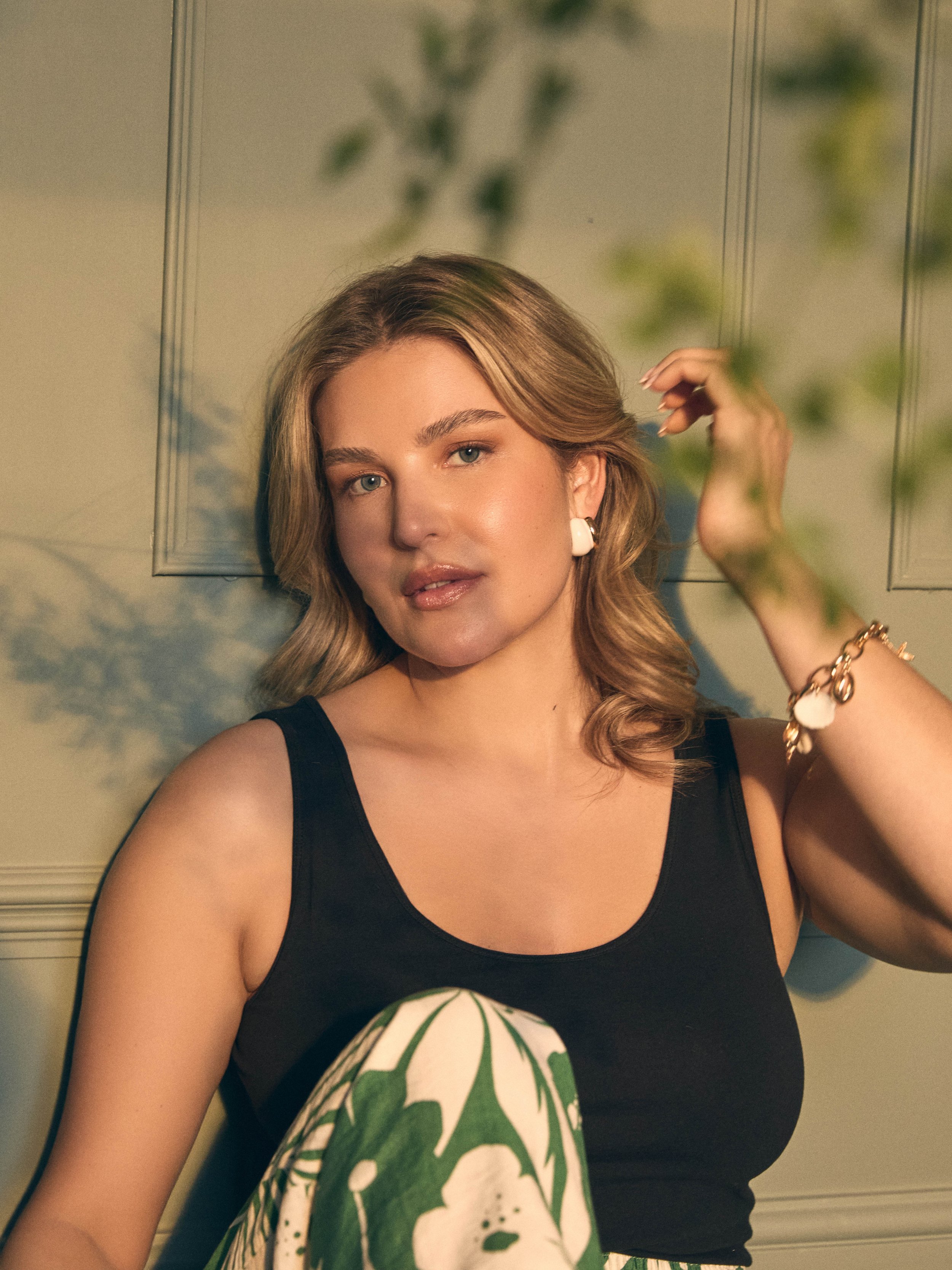 A young woman with shoulder-length blonde hair, wearing a black sleeveless top and patterned pants, sitting against a paneled wall with green shadows from a plant on the wall.