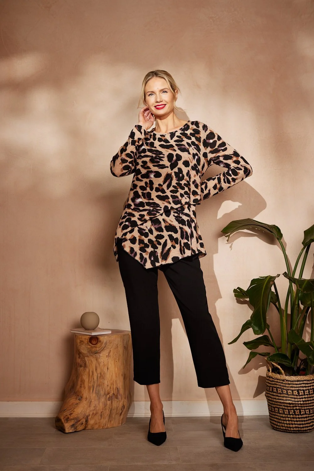 A woman with blonde hair, red lipstick, and a leopard print top standing with one hand on her hip and the other touching her neck in front of a beige wall, a small wooden table with a sphere-shaped object, and a large potted plant.