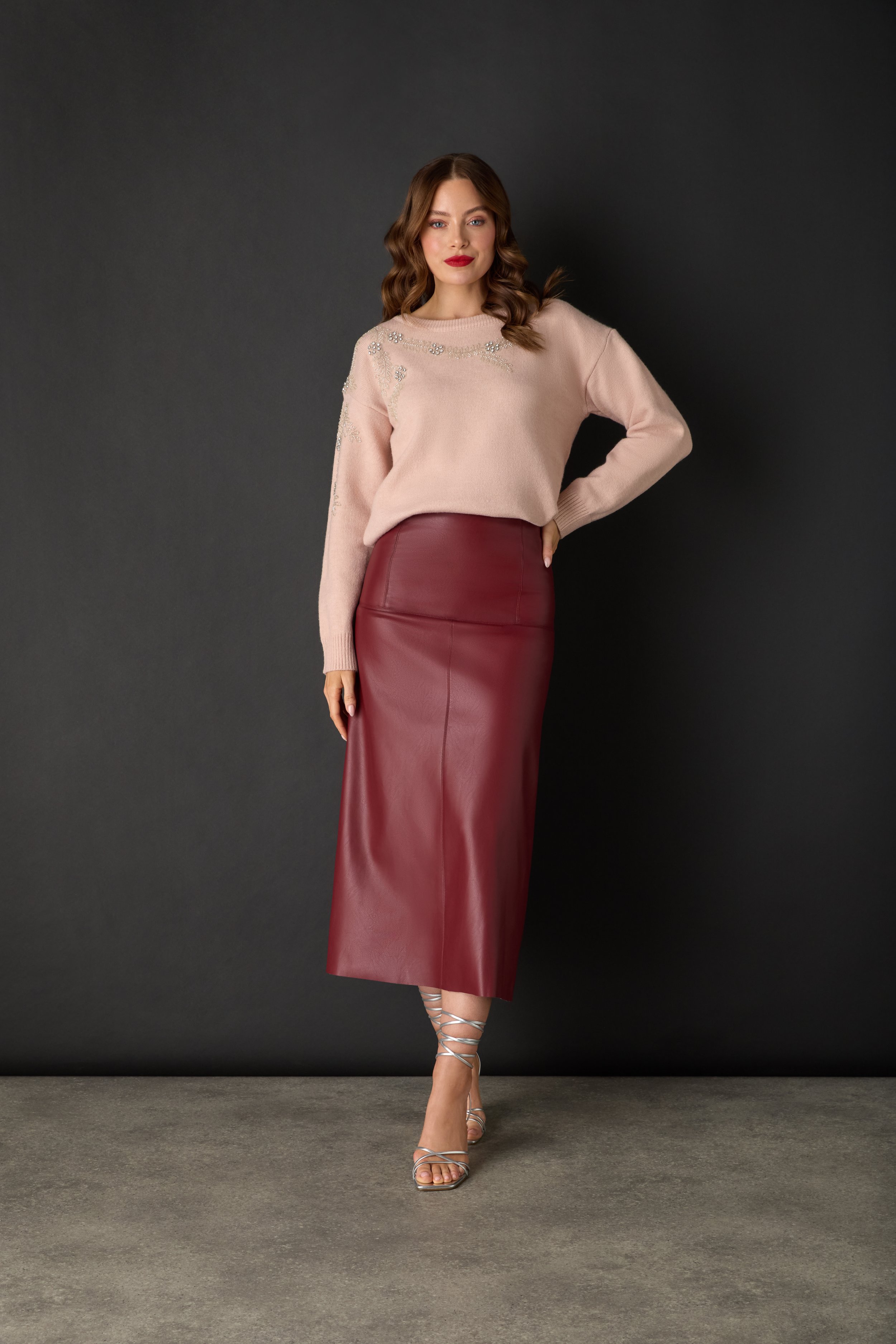 A woman with wavy brown hair wearing a pink sweater with floral embellishments, a high-waisted maroon leather skirt, and silver strappy high heels, standing against a dark gray background.
