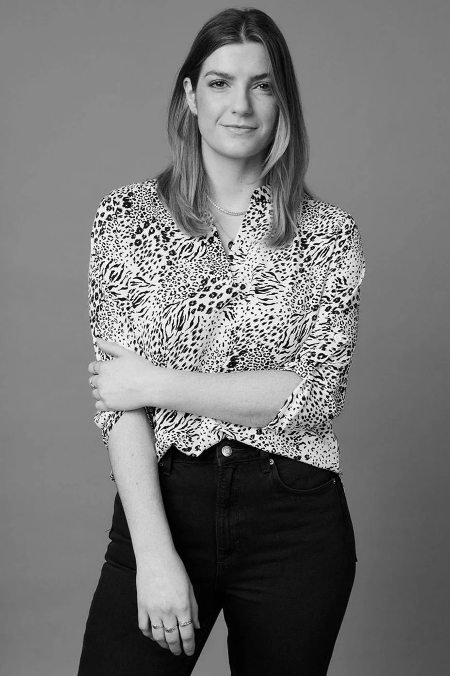 Black-and-white portrait of a woman with shoulder-length hair, wearing a patterned blouse, standing with her arms crossed, looking at the camera with a slight smile against a plain background.