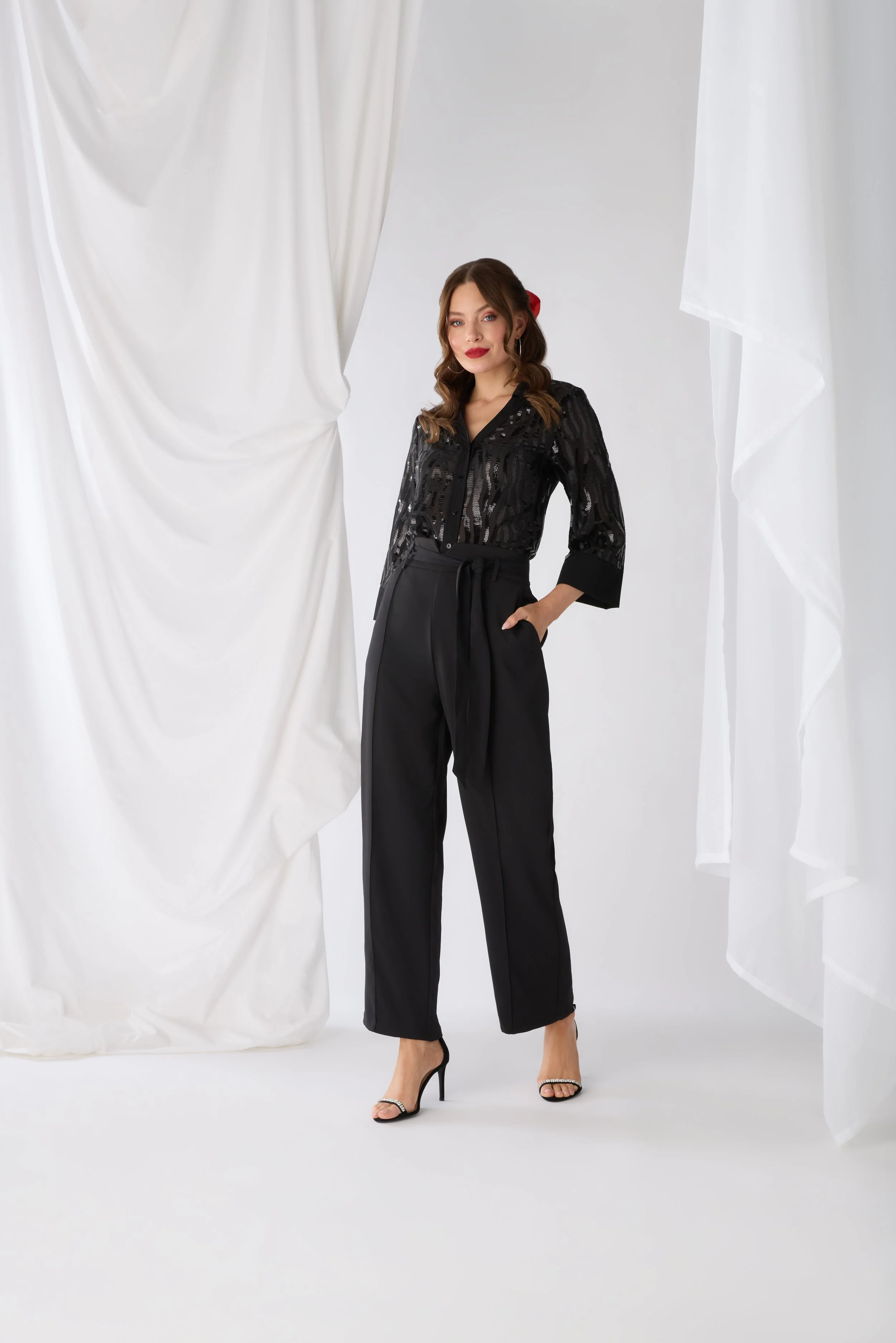 A woman in a black sequined blouse and black high-waisted trousers standing in a studio with white and cream draped curtains.