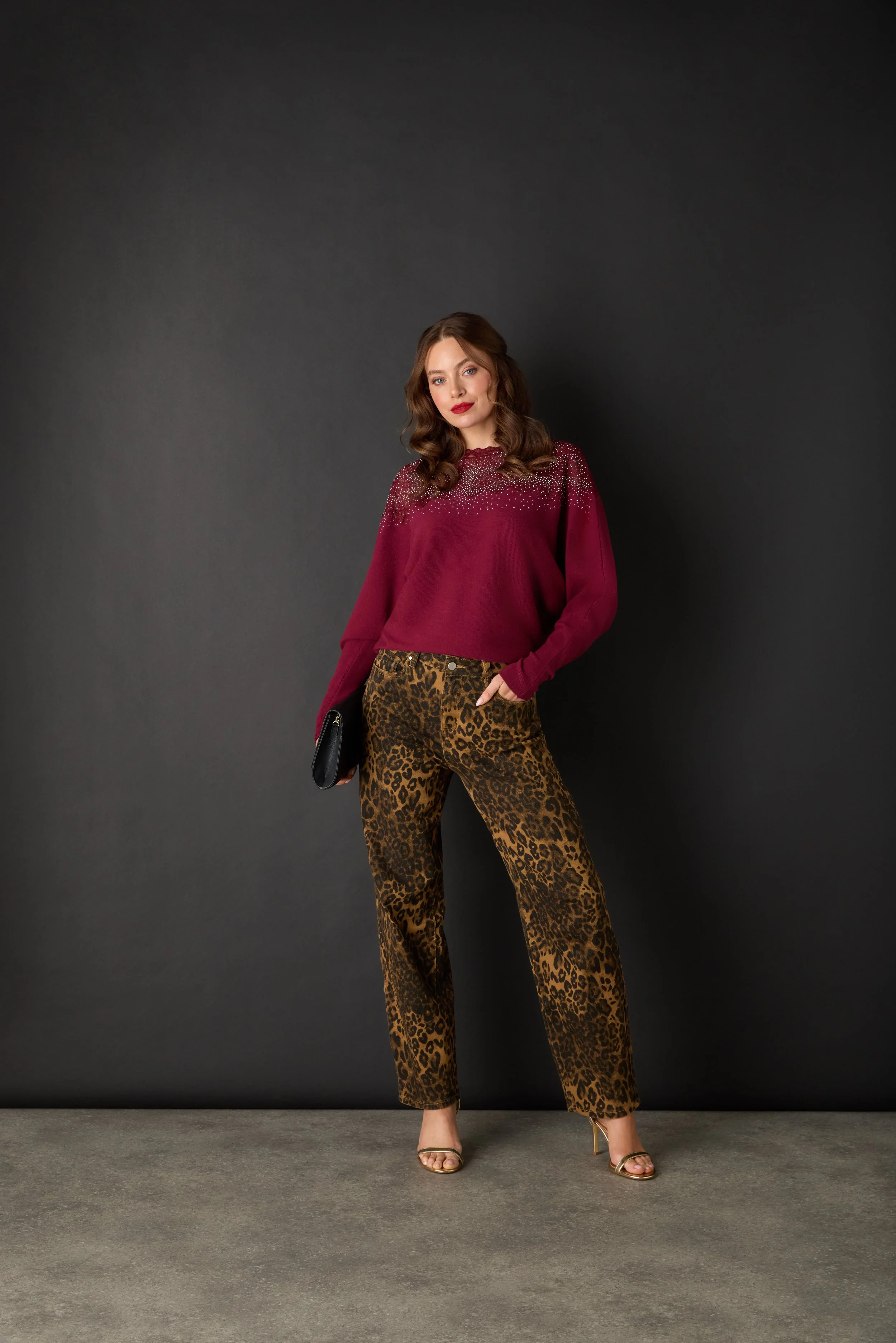 A woman in a red sweater with rhinestone embellishments on the shoulders, leopard print pants, and gold high heels, holding a black clutch, standing against a dark background.