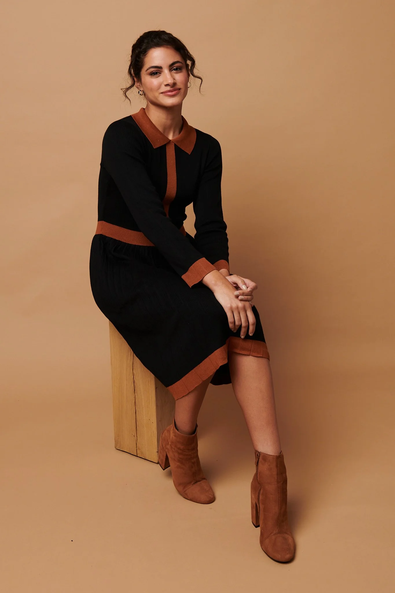 A woman sitting on a wooden block against a beige background, wearing a black dress with brown accents and brown ankle boots.