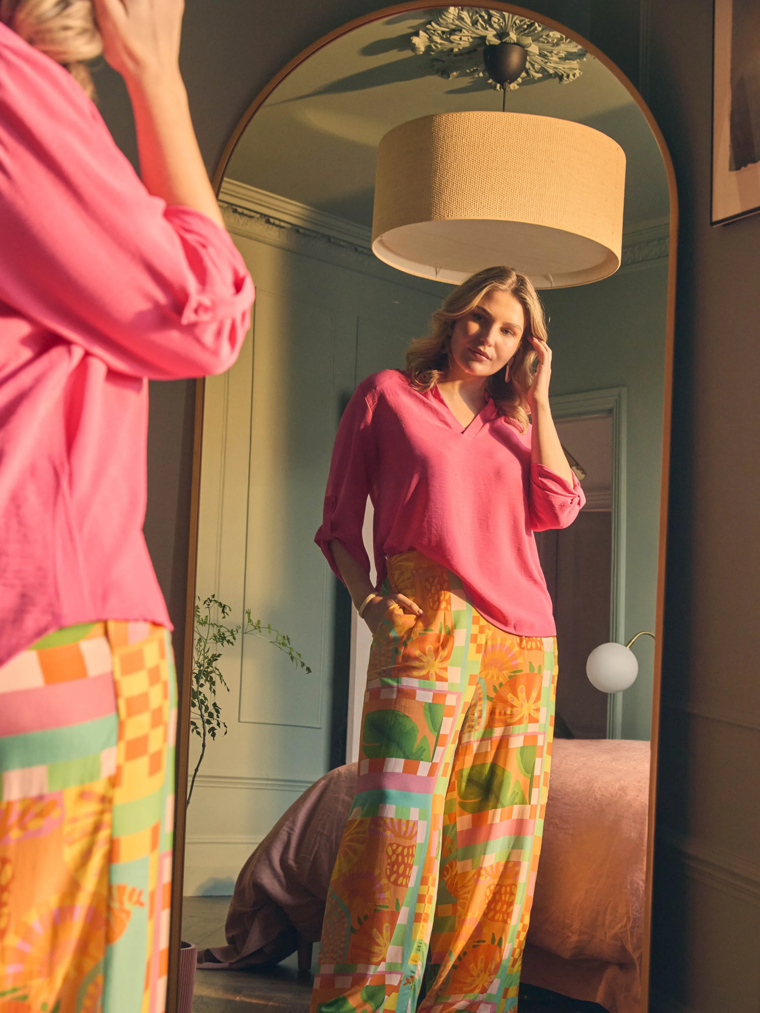 A woman in a pink blouse and colorful patterned pants stands in front of a mirror, adjusting her hair.