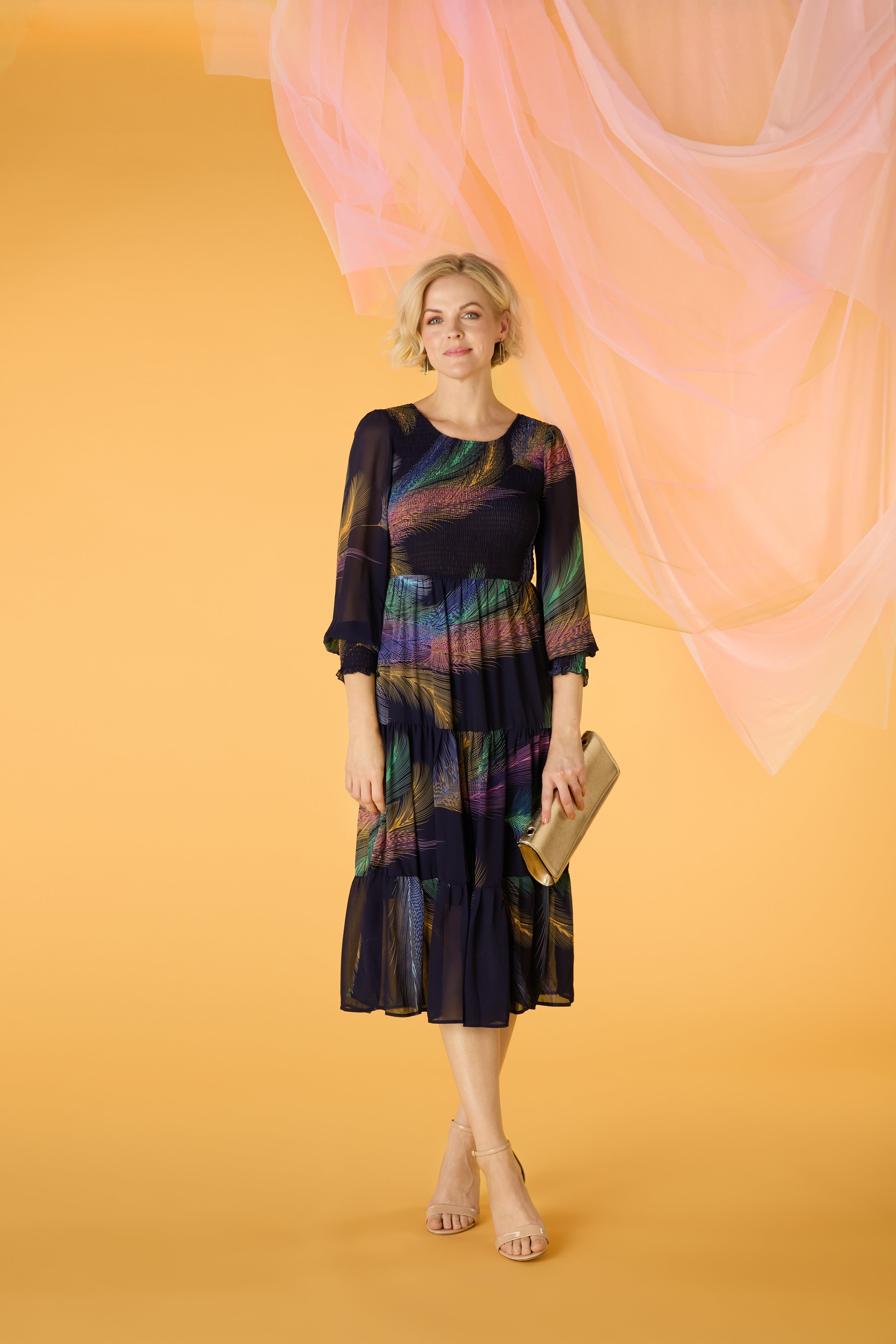 A woman standing against a yellow background with sheer pink fabric draped behind her. She is wearing a black dress with colorful feather-like patterns, nude heels, and holding a beige clutch. She has blonde hair styled in soft waves.