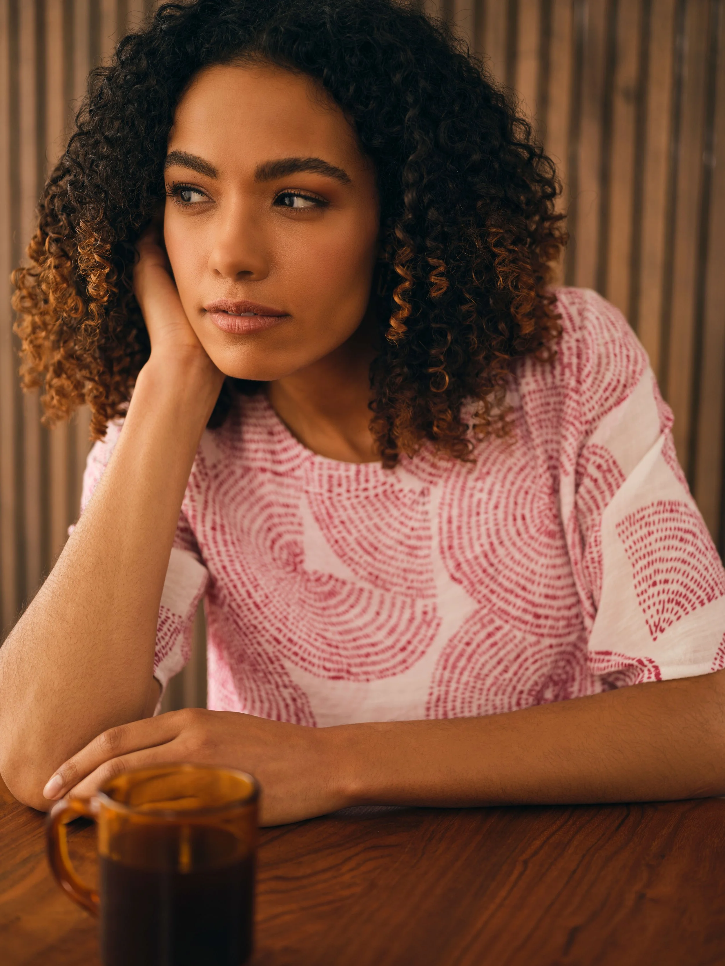 A young woman with curly hair sits at a wooden table with a mug of coffee in front of her, resting her head on her hand and looking pensively to the side.