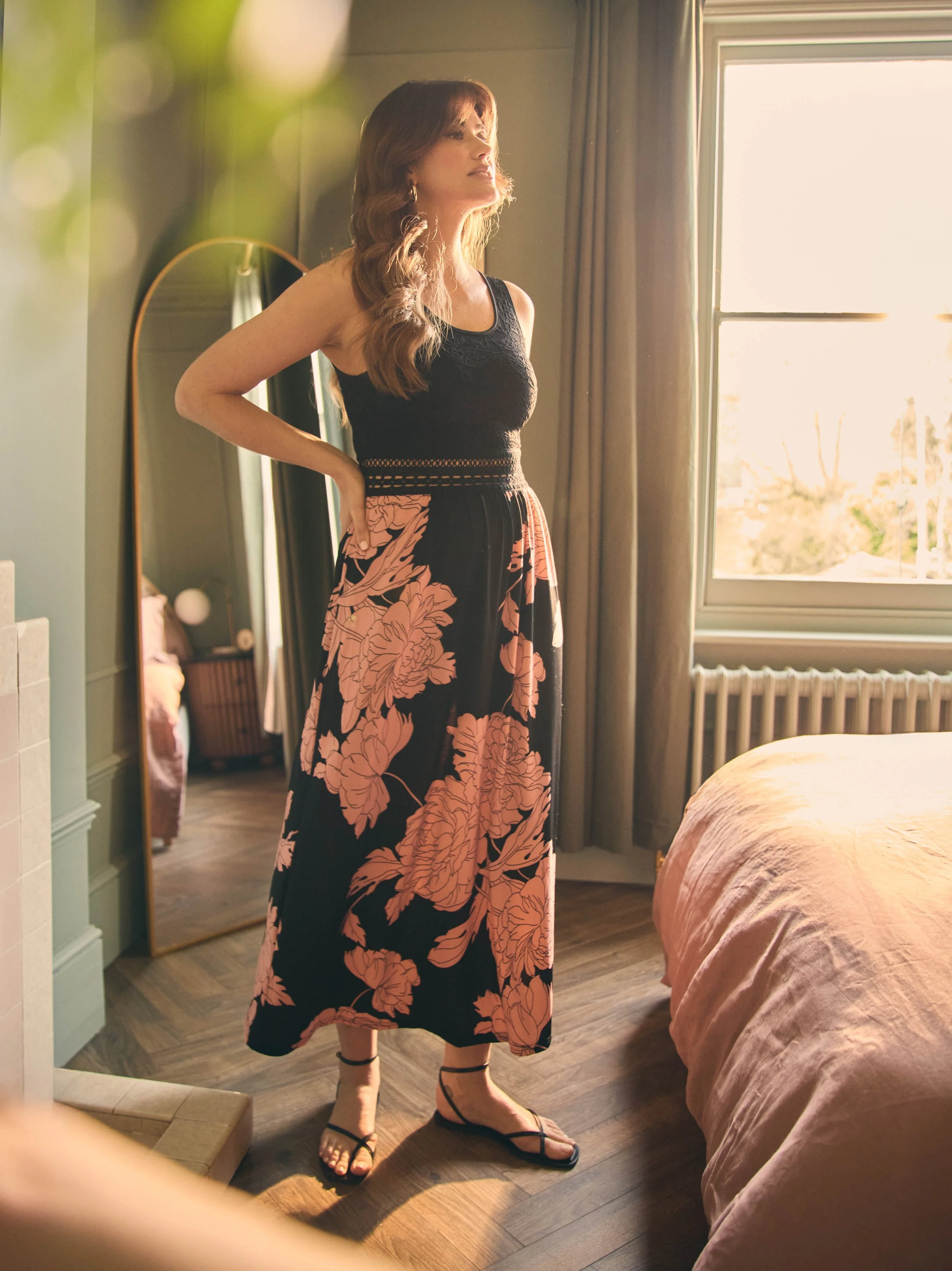 A woman with long, wavy brown hair standing in a sunlit bedroom, wearing a black sleeveless top and a long floral skirt with pink flowers, black sandals, looking out the window.