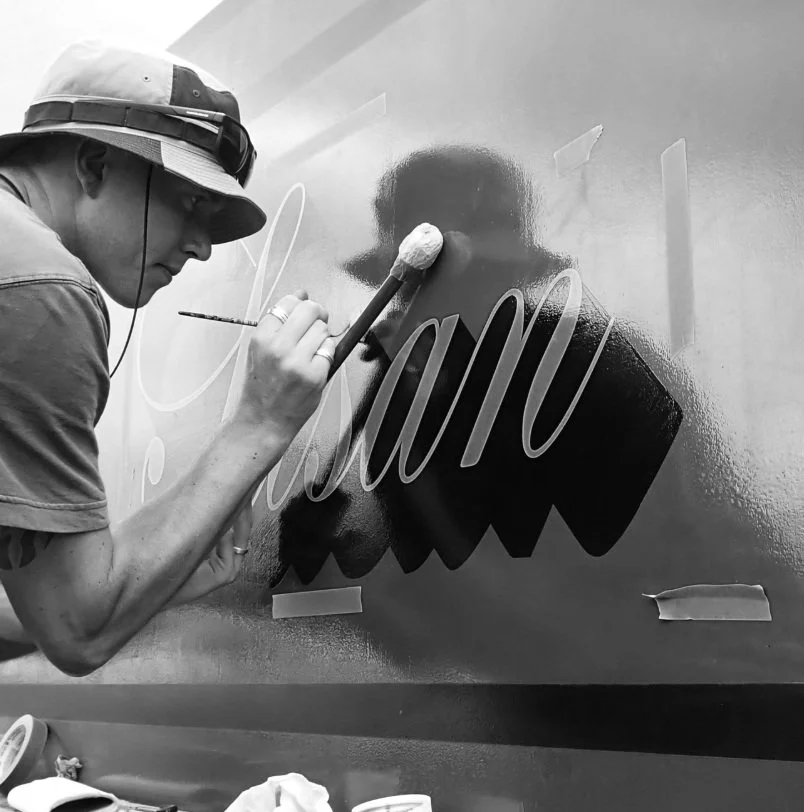 A person wearing a hat and glasses is painting a sign on a wall with a brush, creating cursive lettering.