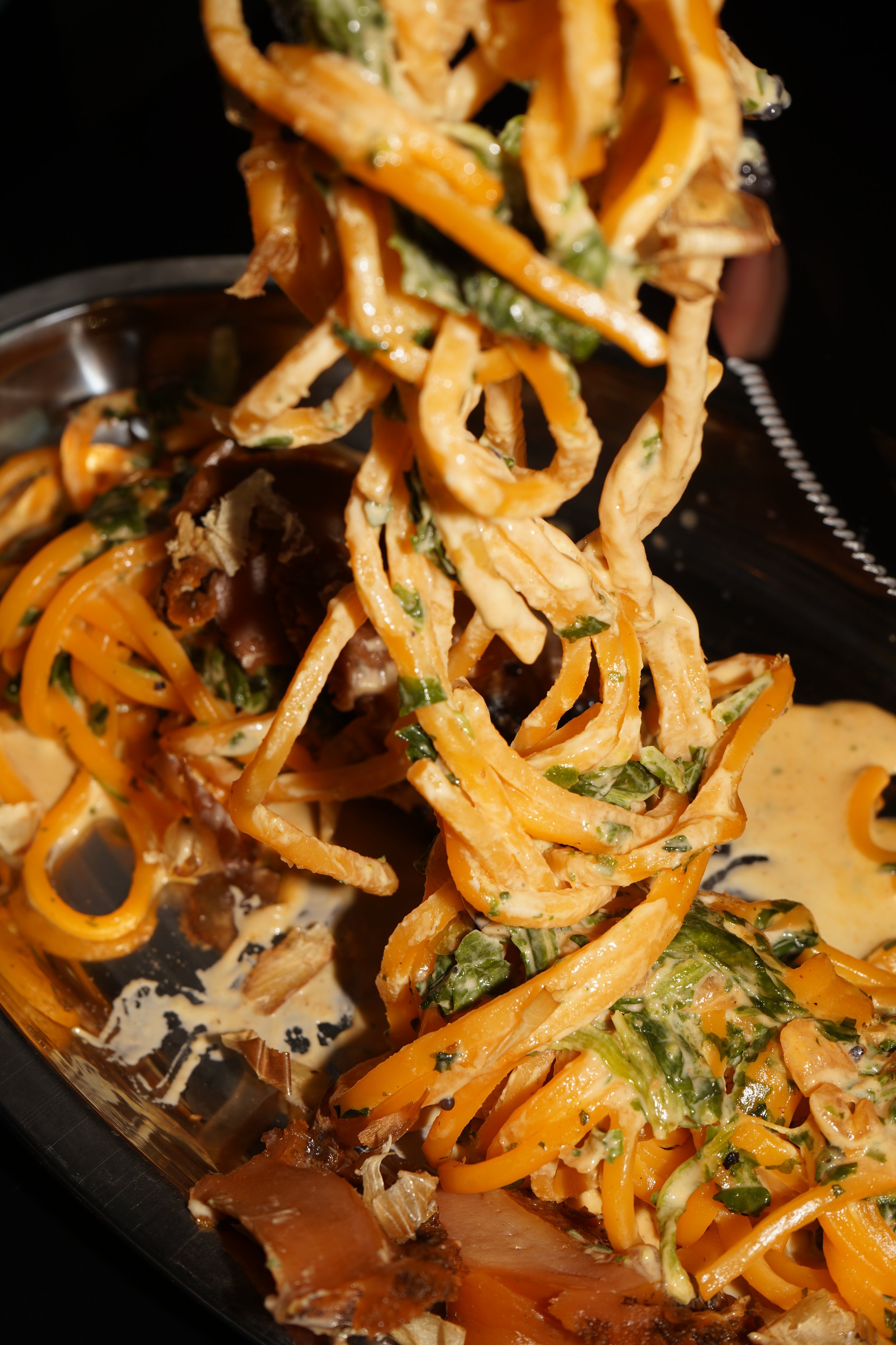 Close-up of a serving of stir-fried noodles with vegetables and sauce being lifted with chopsticks on a black plate.