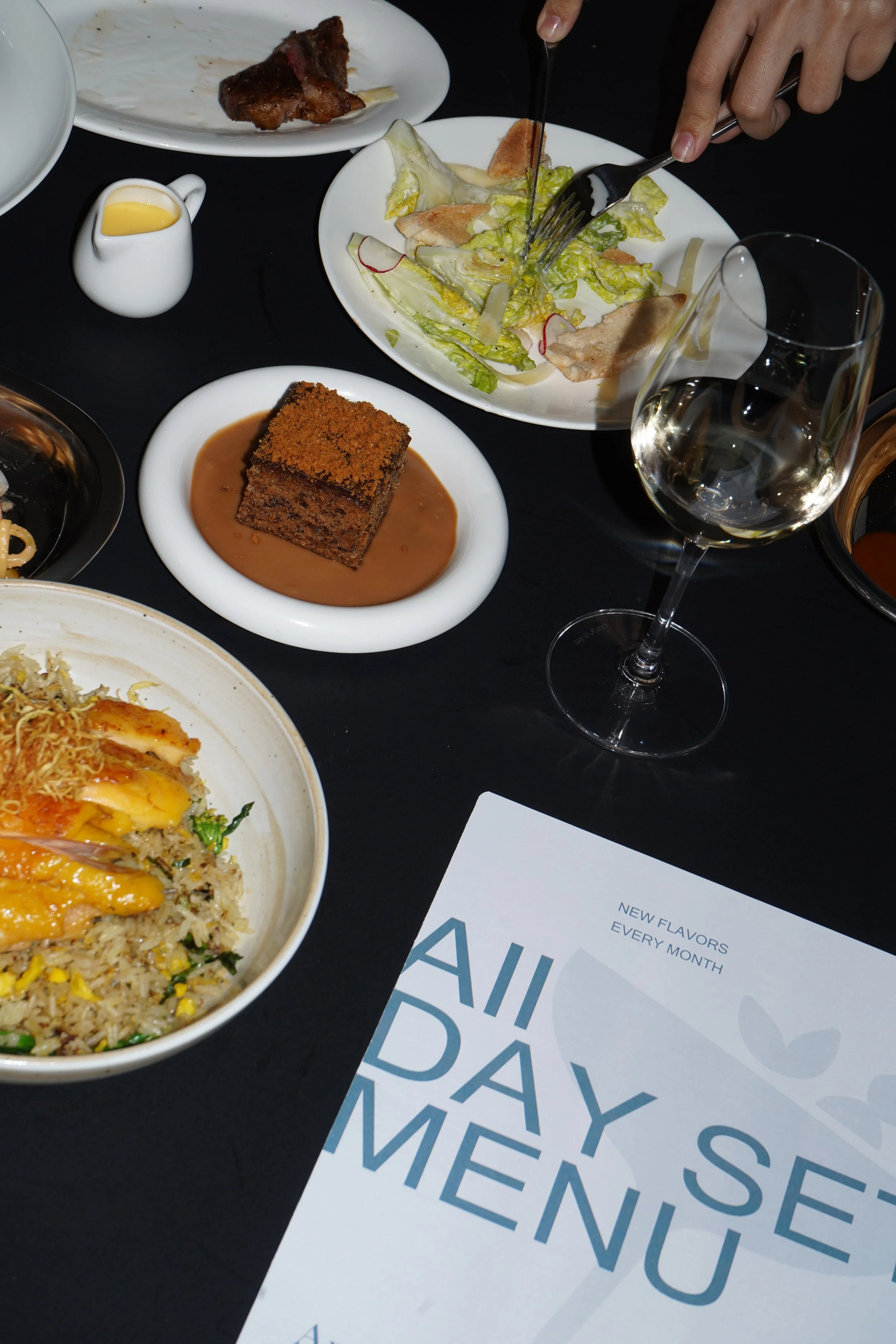 Diverse dishes on a table, including a bowl of fried rice with sliced chicken, a piece of cake in sauce, salad with greens and radish slices, a glass of white wine, and a small pitcher of dressing. Part of a menu or brochure is also visible.