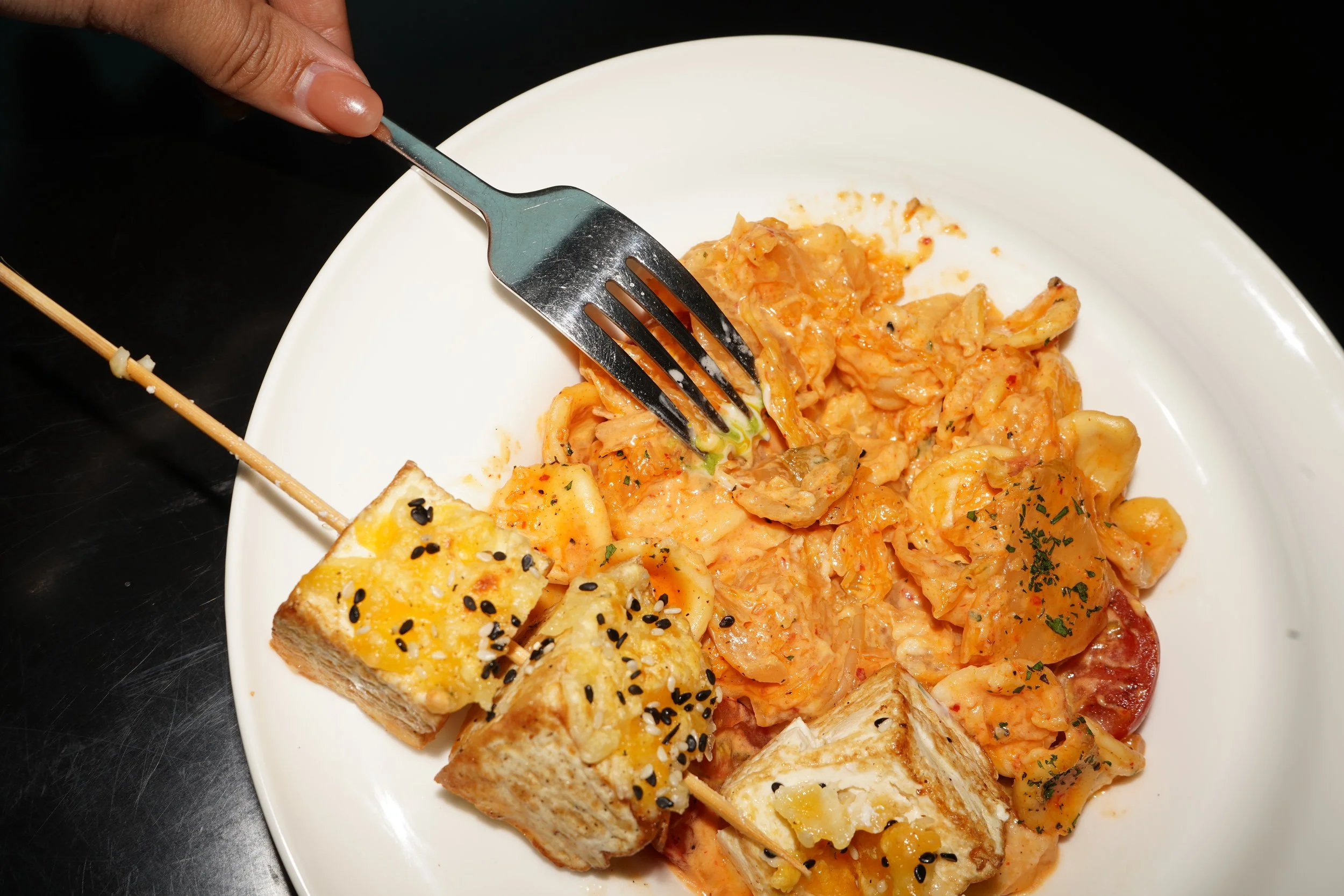A white plate of saucy, cooked chicken topped with herbs, and two pieces of toast with melted cheese and black sesame seeds, served with a fork and skewer on a black surface.