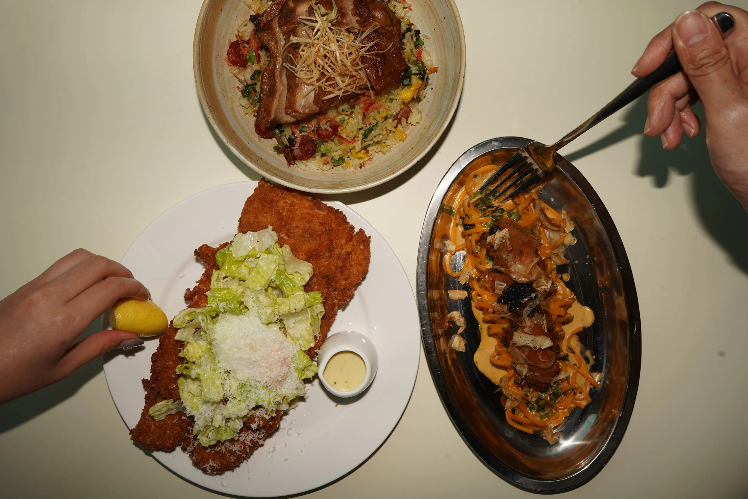Top-down view of a table with three plates of food. The top plate has rice mixed with vegetables and topped with cooked meat and shredded cheese. The middle plate contains a breaded fried fish fillet with a side of chopped lettuce and a small container of creamy dressing, with a lemon wedge being held above by a hand. The right side has an oval-shaped metal dish with pasta topped with shredded meat and sauce, and a hand holding a fork.