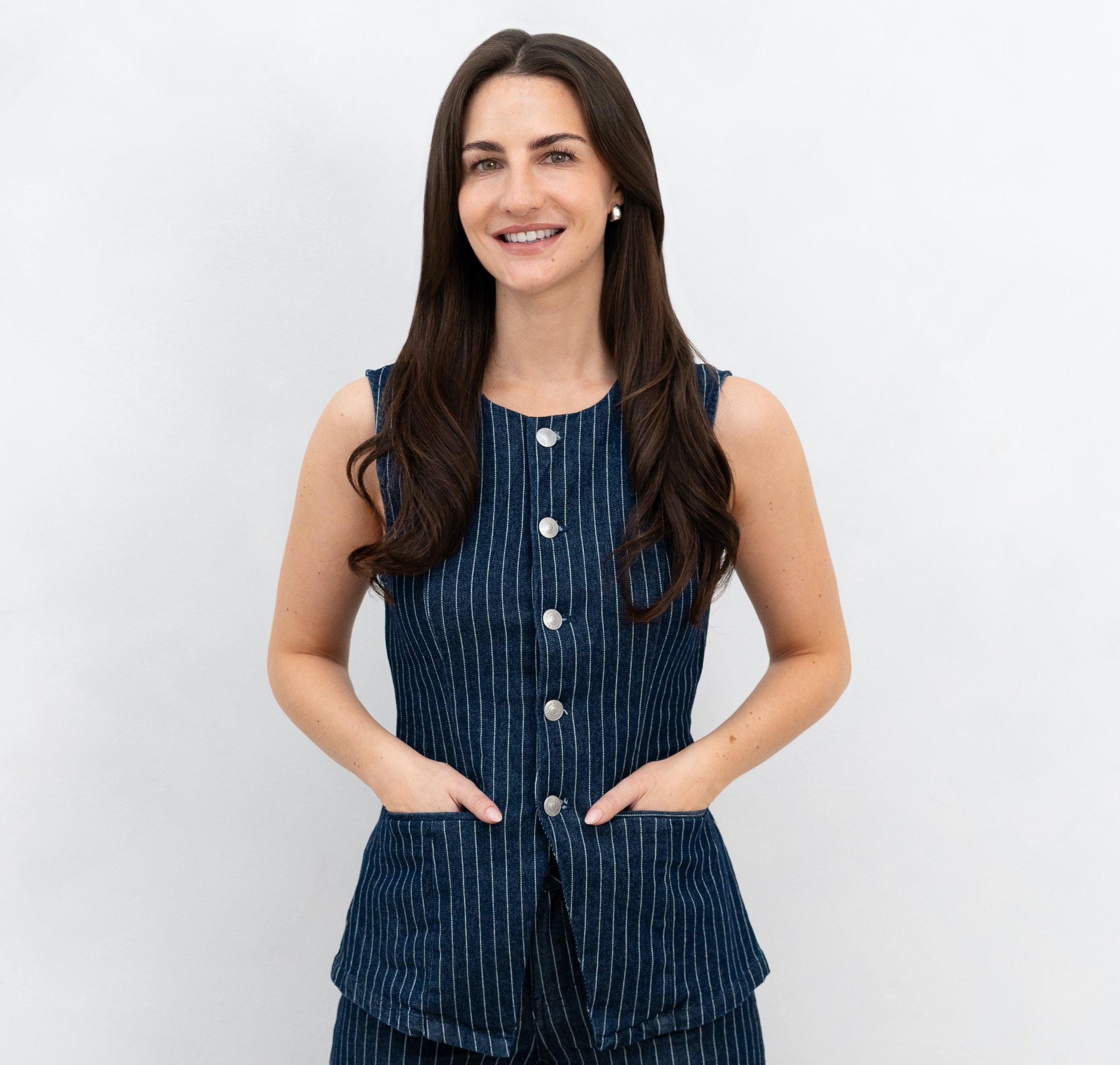 A young woman with long brown hair, wearing a sleeveless denim dress with vertical stripes and buttons, standing against a plain white background.