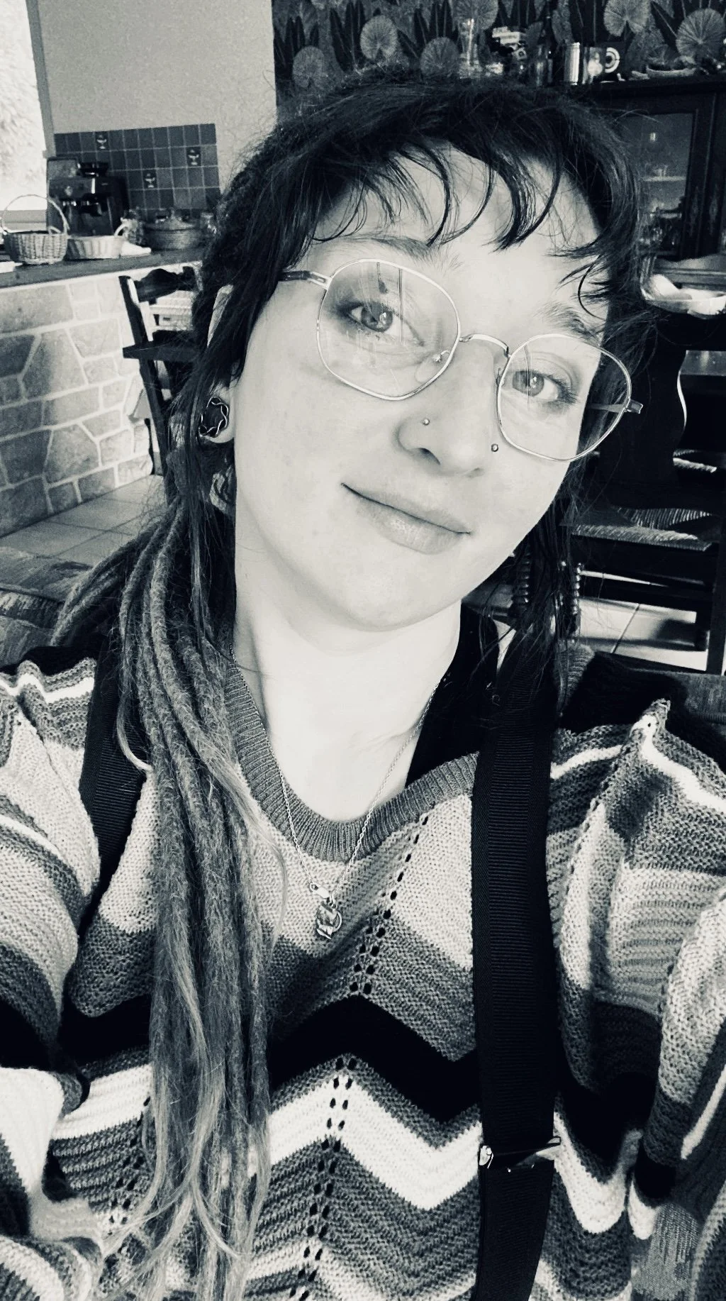 Un portrait en noir et blanc d'une jeune femme avec des lunettes rondes, des dreadlocks et un sweat à motifs. Elle porte un collier avec un pendentif et un piercing au nez.