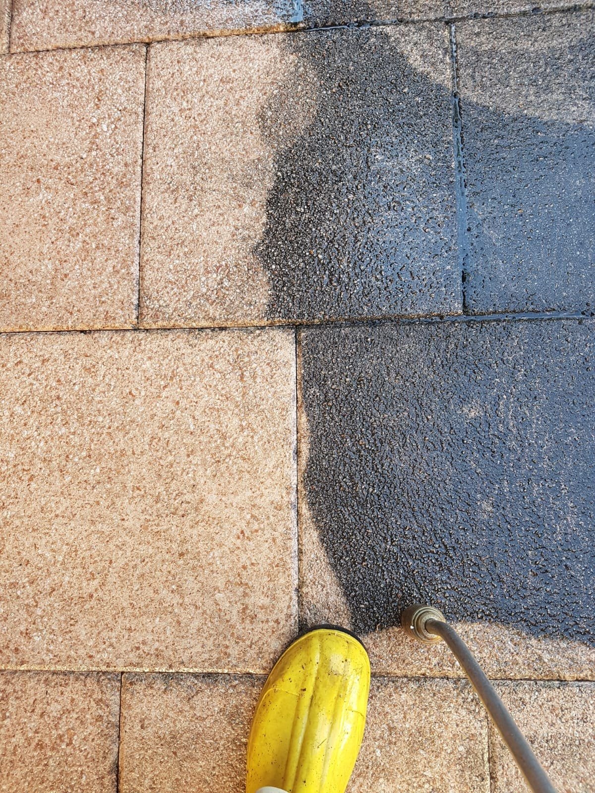 Ein Gehweg wird mit einem Hochdruckreiniger gereinigt, wobei der Unterschied zwischen sauberer und verschmutzter Fläche sichtbar ist. Gelber Gummistiefel im Vordergrund.