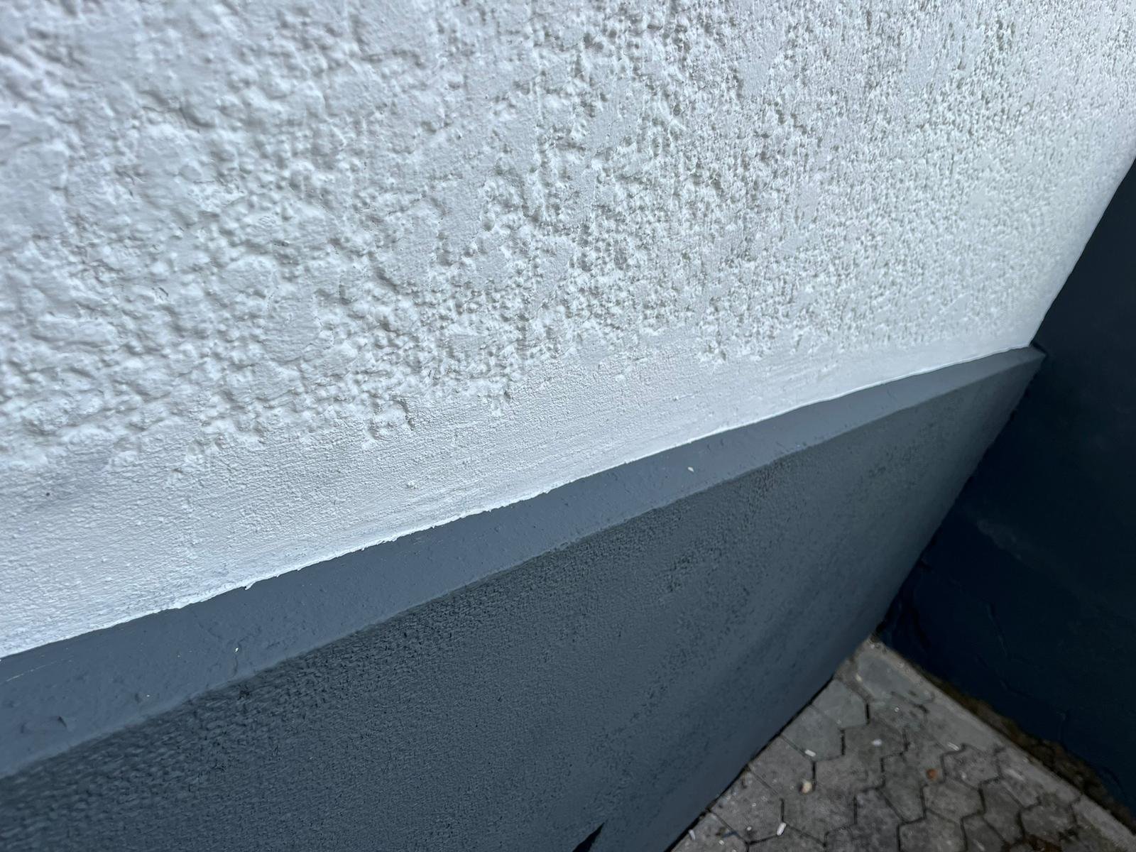 Nahaufnahme einer Hauswand mit rauer weißer Struktur, einem grauen Sockel und Kopfsteinpflasterboden.