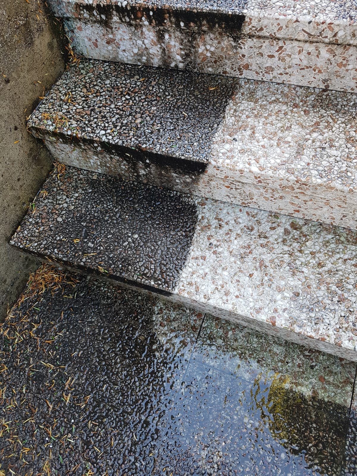 Naheaufnahme von steinernen Stufen, die teilweise durch Wasser oder Schmutz dunkel verfärbt sind, mit deutlichem Kontrast zwischen sauberem und verunreinigtem Bereich. Regenpfützen und Schmutz sind sichtbar.