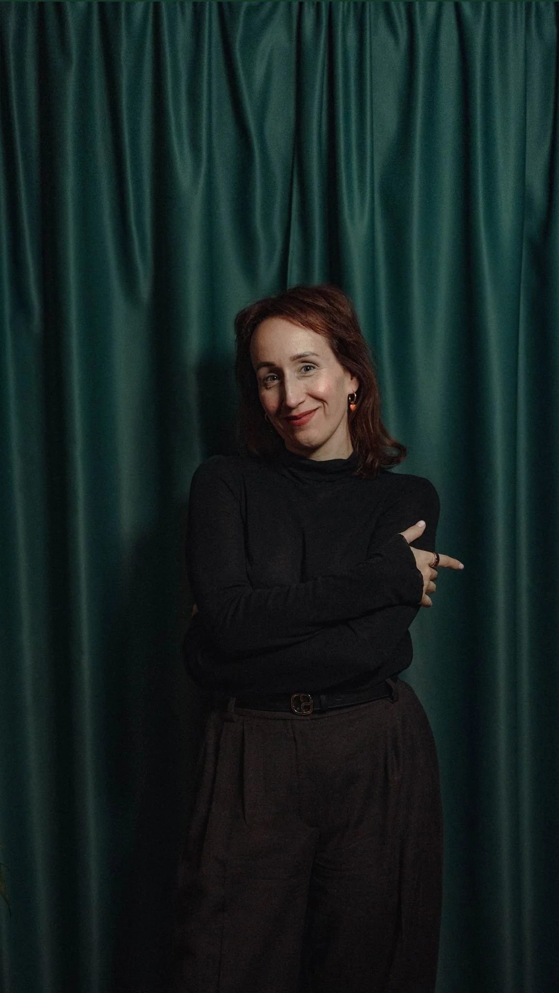 Person in dark clothing standing in front of dark green curtains, smiling.