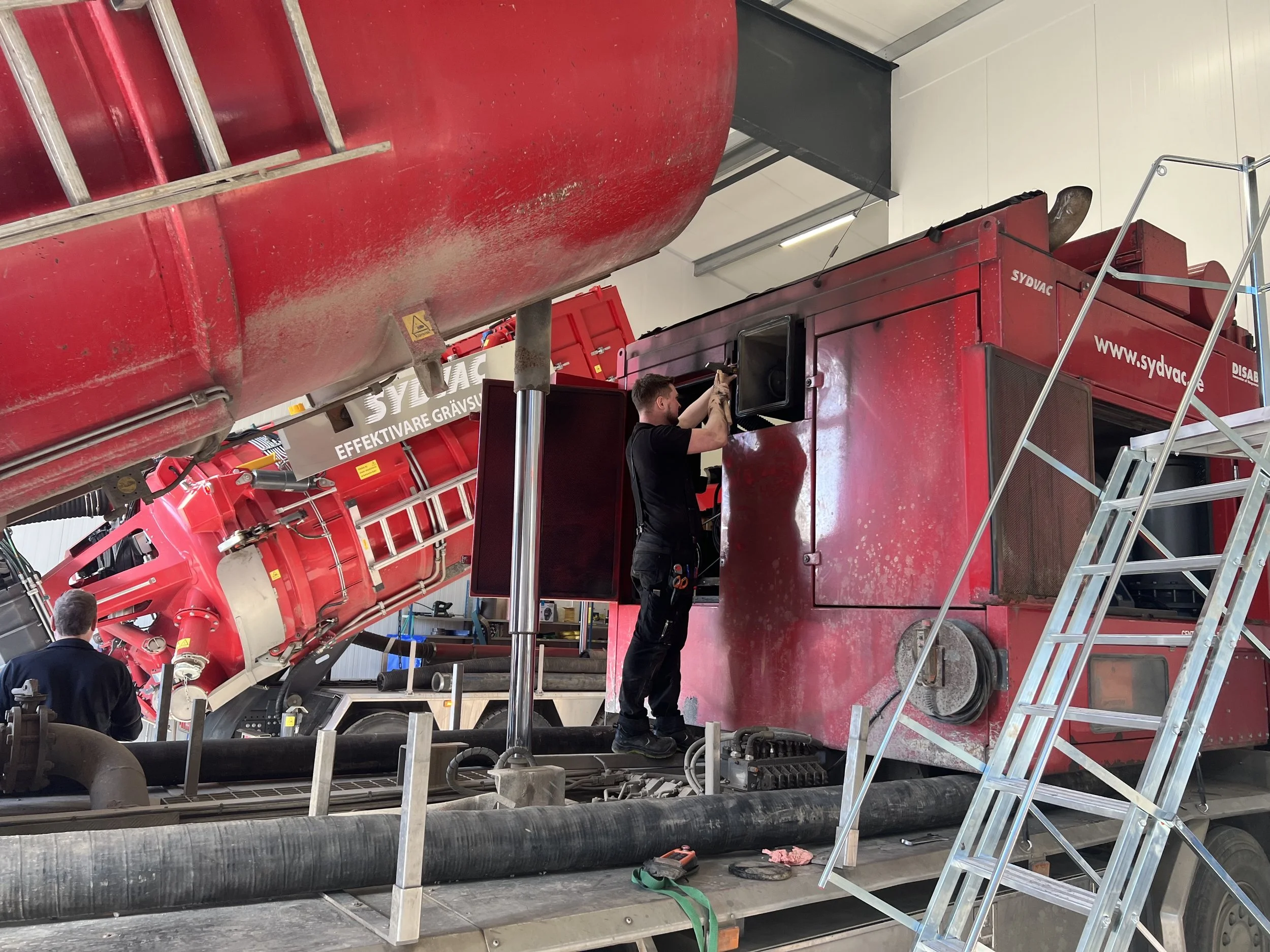 En person arbetar med en stor röd vakuumlastare i en verkstadsmiljö. Det finns stegar och slangar runt omkring maskinen.