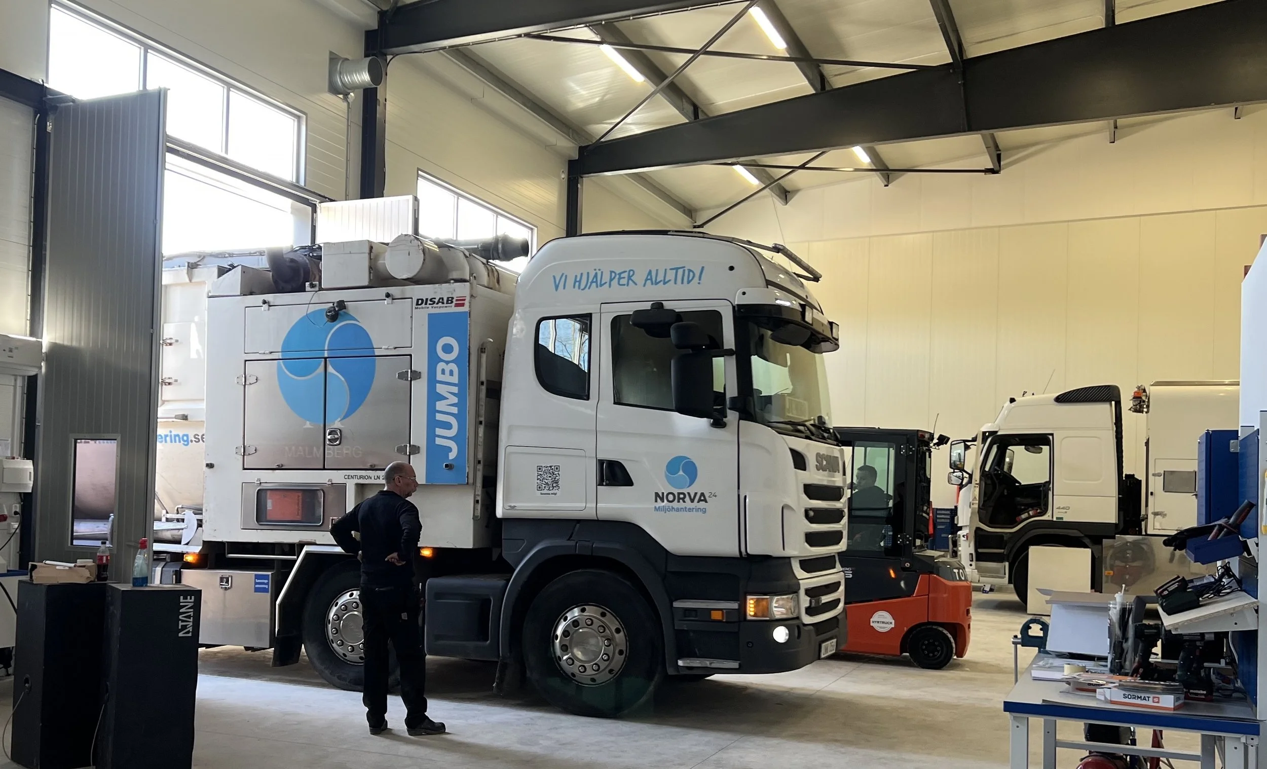 Slamsug i ett garage med texten 'Vi hjälper alltid!' och företagslogotyper, omgiven av arbetsutrustning och ett bord med verktyg.