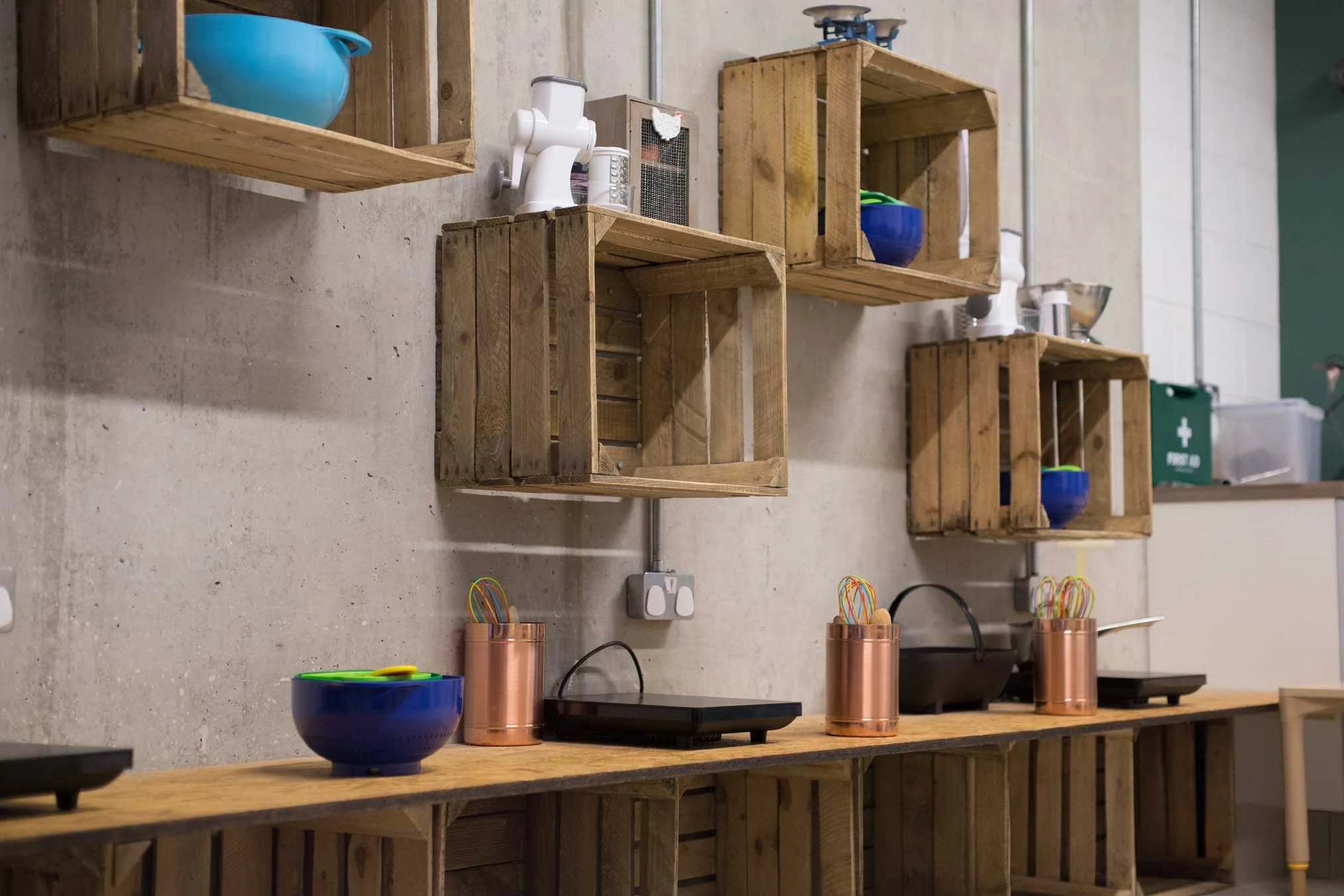 Wooden crates mounted on a concrete wall, containing kitchen items, with copper containers and children's toy appliances on a wooden counter simulating a kitchen.