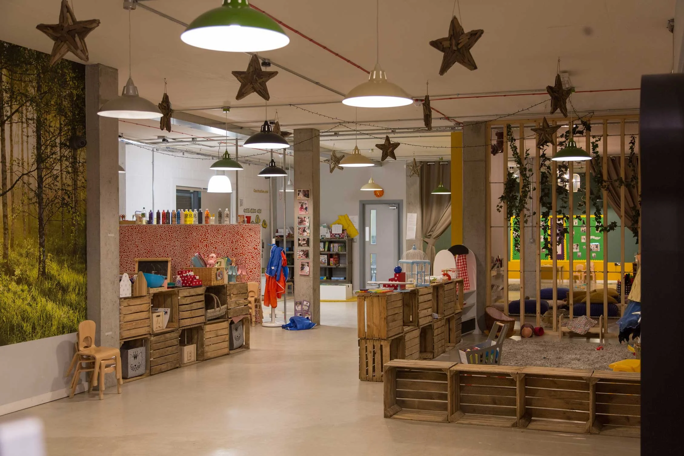 Interior of a children’s playroom with hanging star decorations, wooden crates, small chairs, and various toys and books, with a mural of a forest scene on the wall.