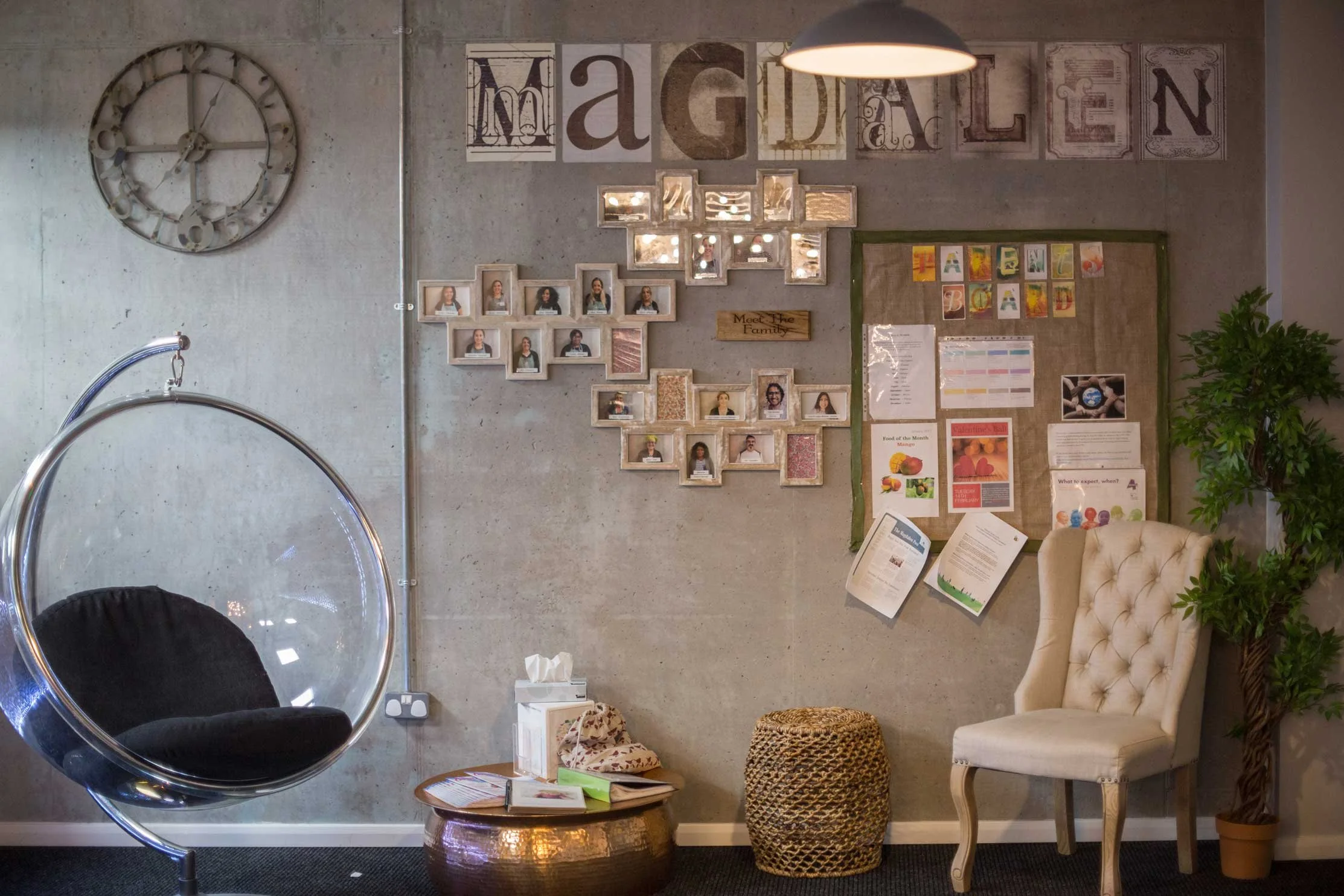 Reception area at The Grove with a large clock on a concrete wall, picture frames, a bulletin board, a white upholstered chair, a hanging bubble chair with black cushion, a round metallic table, a wicker stool, tissue box, books, and an indoor plant.