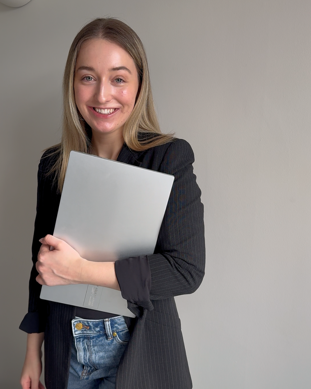 A young woman with long blonde hair smiling and holding a silver laptop, wearing a black blazer and jeans, standing against a plain light-colored wall.