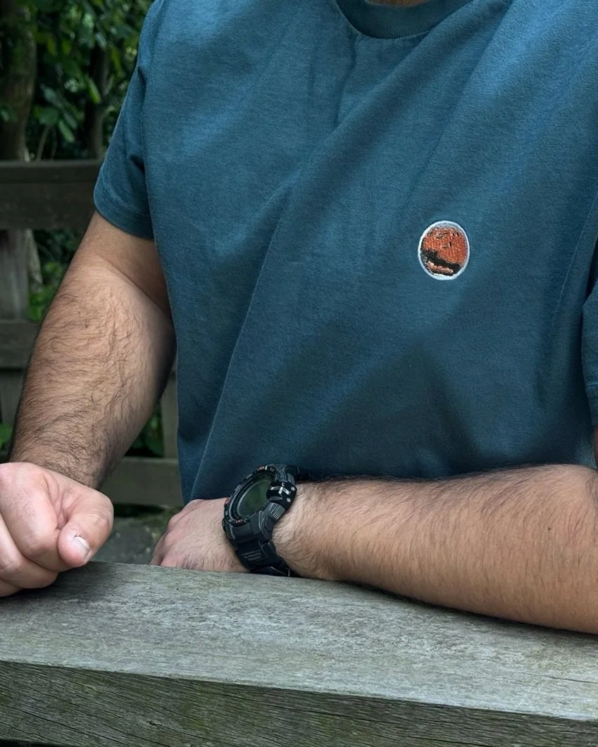 A person wearing a dark blue t-shirt with a small round embroidered design of Mars, sitting outdoors at a wooden surface, wearing a black digital watch on their left wrist, with a background of green foliage.