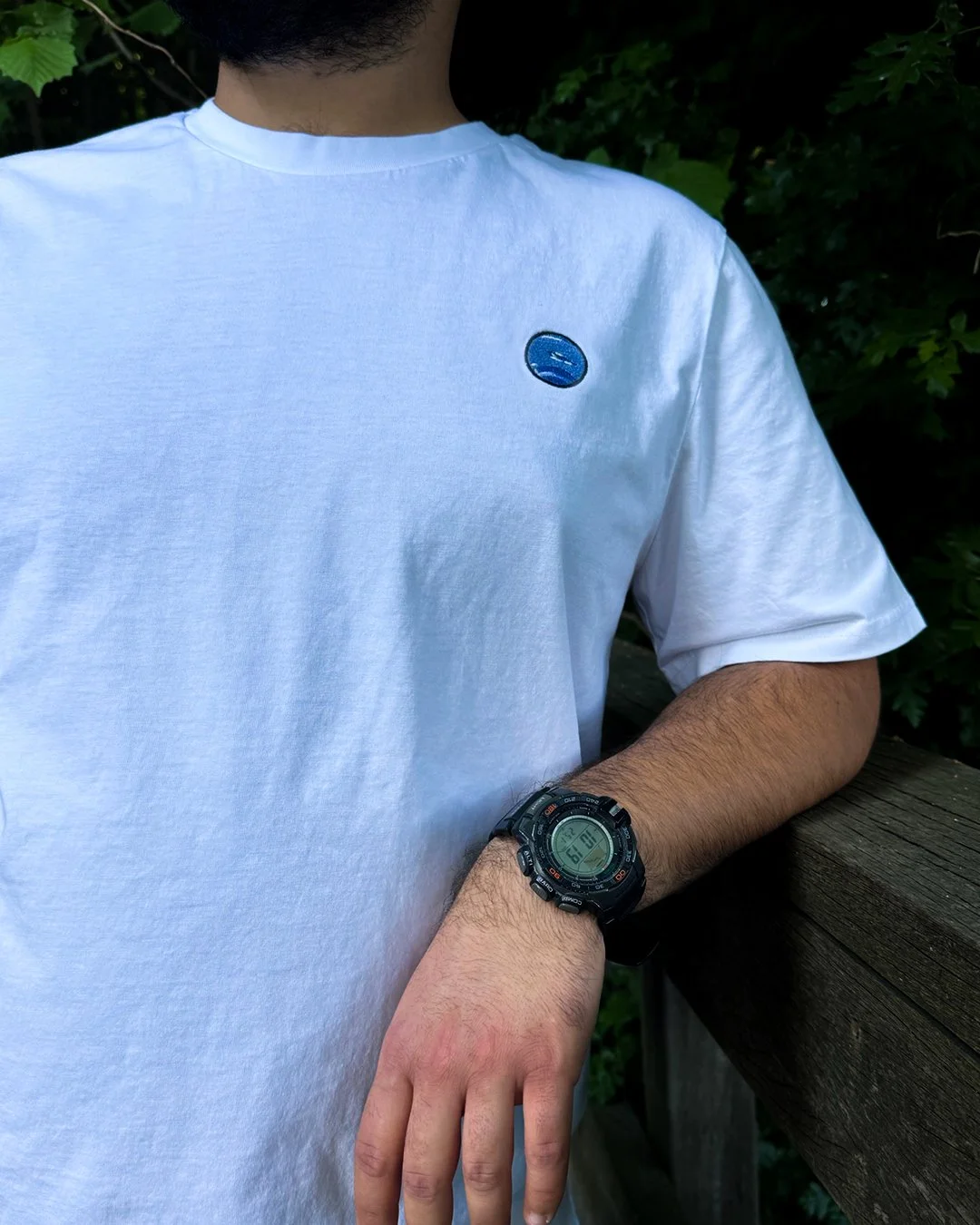 Person wearing a white t-shirt with a small blue embroidered logo on the chest and a black digital wristwatch, standing outdoors next to a wooden railing with green foliage in the background.