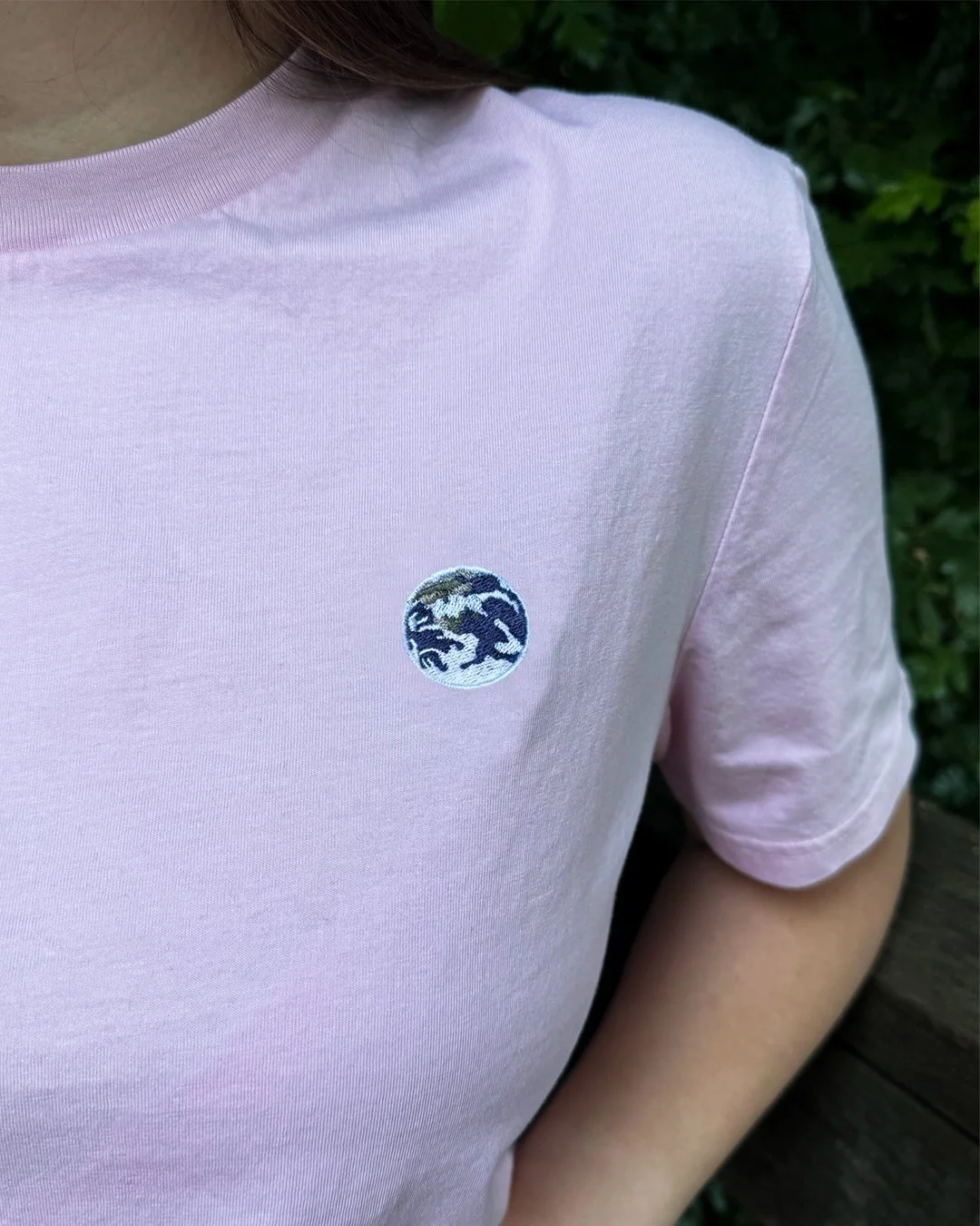 Close-up of a person wearing a light pink T-shirt with a small embroidered patch of a Earth on the chest, outdoors with green foliage in the background.