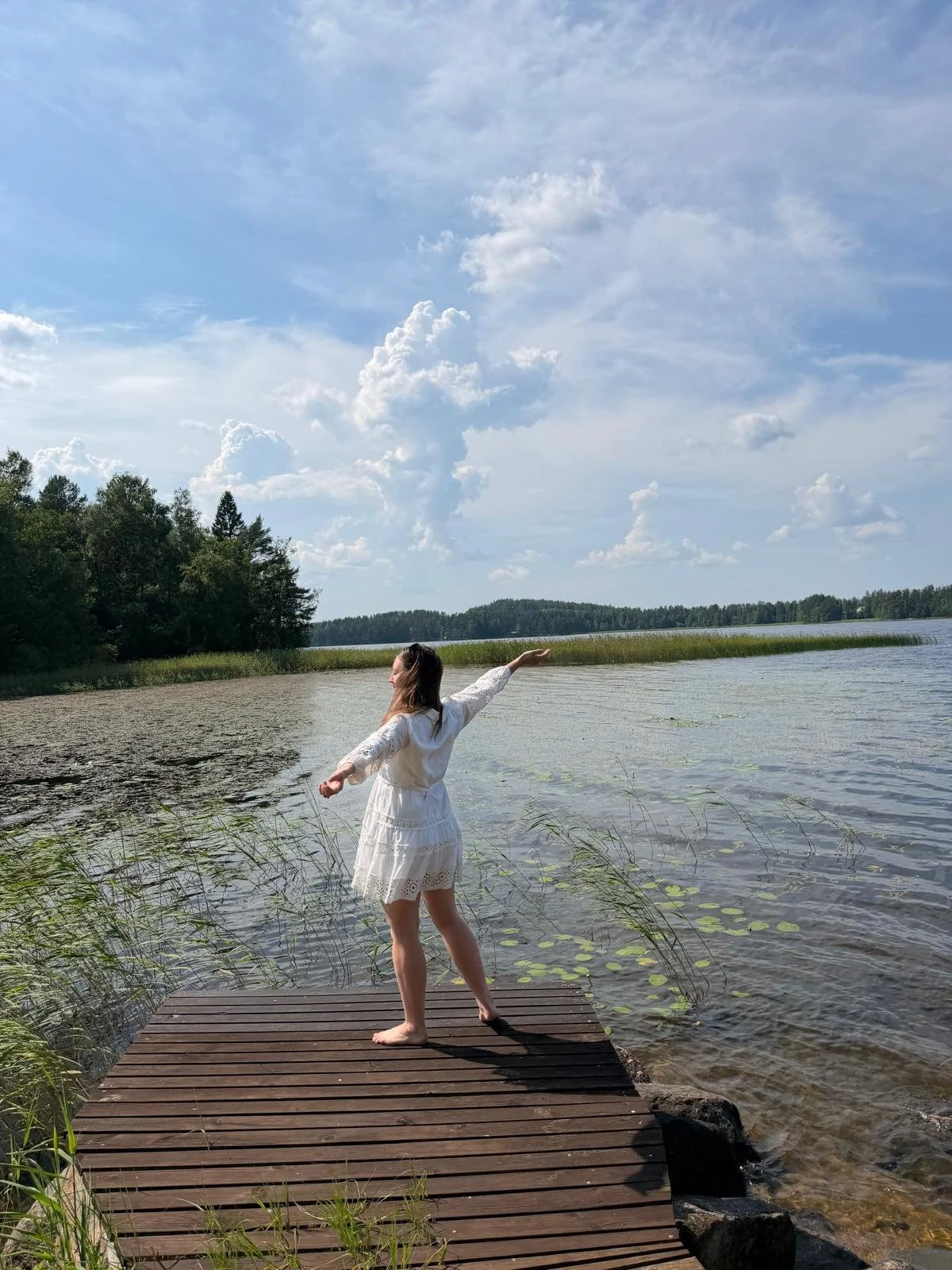 Roya stands on a wooden pier overlooking the calm waters of Lake Saimaa, her arms open wide in a gesture of freedom and joy after her Finnish Happiness Camp experience at Blueberry Villa.