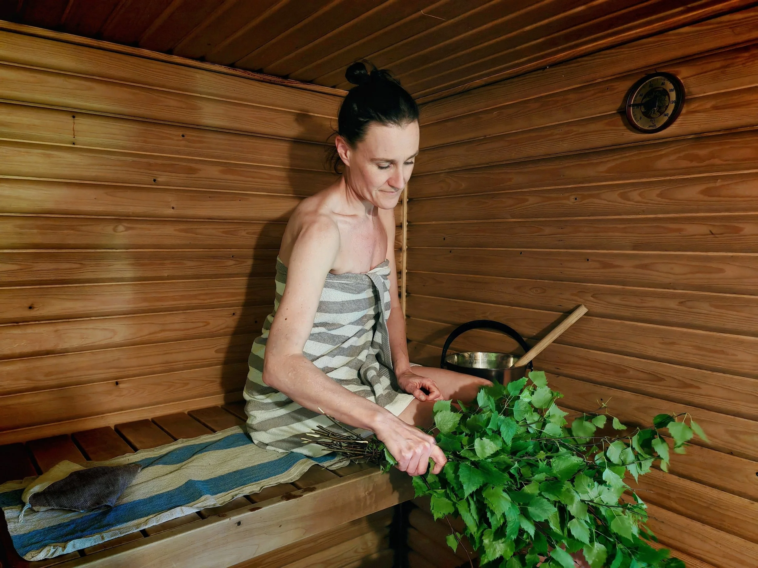 A woman relaxes in the traditional lakeside sauna at Blueberry Villa, holding a birch whisk made from branches gathered in the villa's own garden. Surrounded by the warmth of the wooden sauna, she embraces the slow rhythm of Finnish sauna culture.