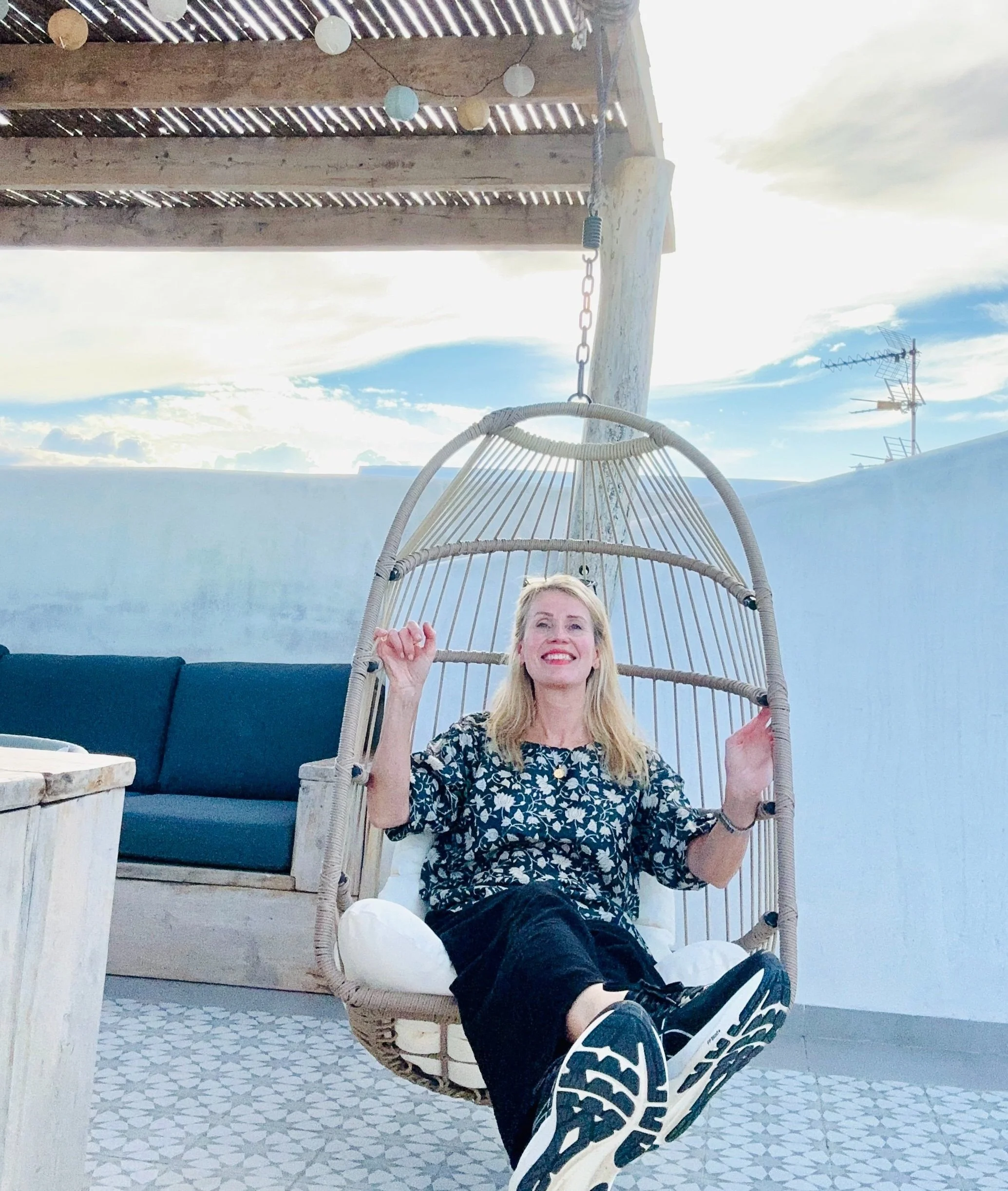 A smiling transformative travel coach Mirka Kristiina sitting in a hanging chair on a rooftop patio with a blue sky and white wall visible in the background.