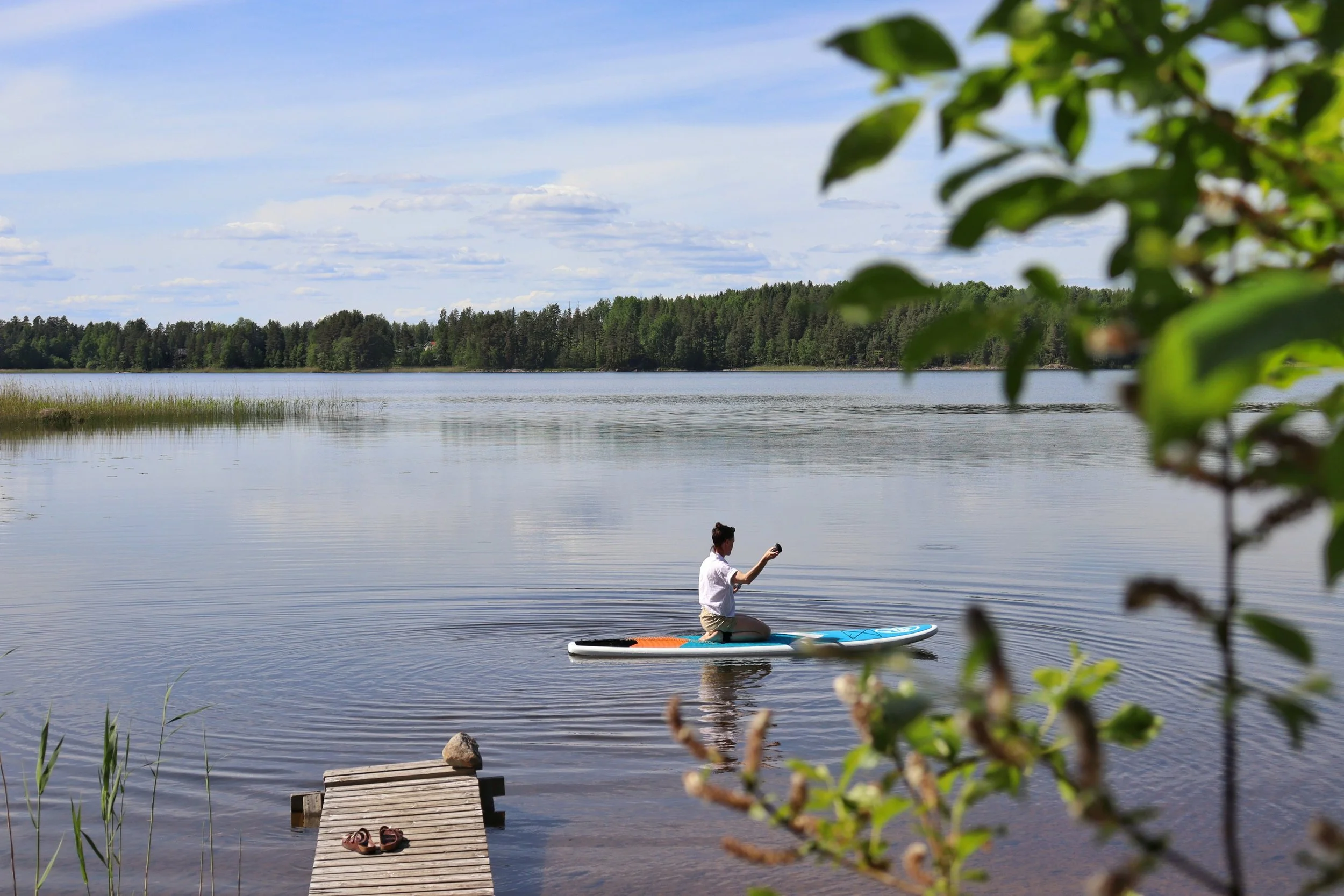 A stand-up paddleboard rests on the shore of Blueberry Villa on Lake Saimaa, inviting guests to explore the calm, crystal-clear waters surrounded by Finnish forest and peaceful lakeside scenery. There is also a rowing boat and fishing equipment.