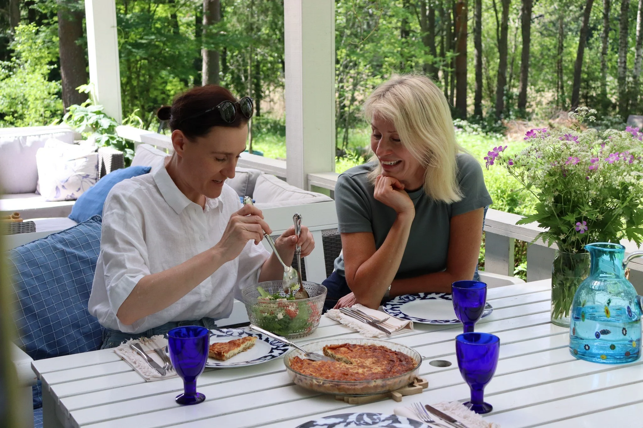 Two women enjoy a sunny afternoon lunch on the terrace at Blueberry Villa, sharing a homemade quiche made with chanterelle mushrooms handpicked from the surrounding forest. The relaxed meal celebrates seasonal Finnish flavors and the peace.