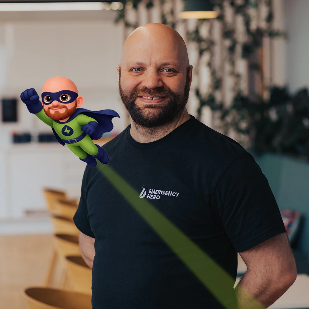 A man with a beard wearing a black t-shirt with 'Emergency Hero' logo, standing indoors, with a cartoon superhero figure of a bald man with a beard, glasses, green suit, and cape floating beside him.