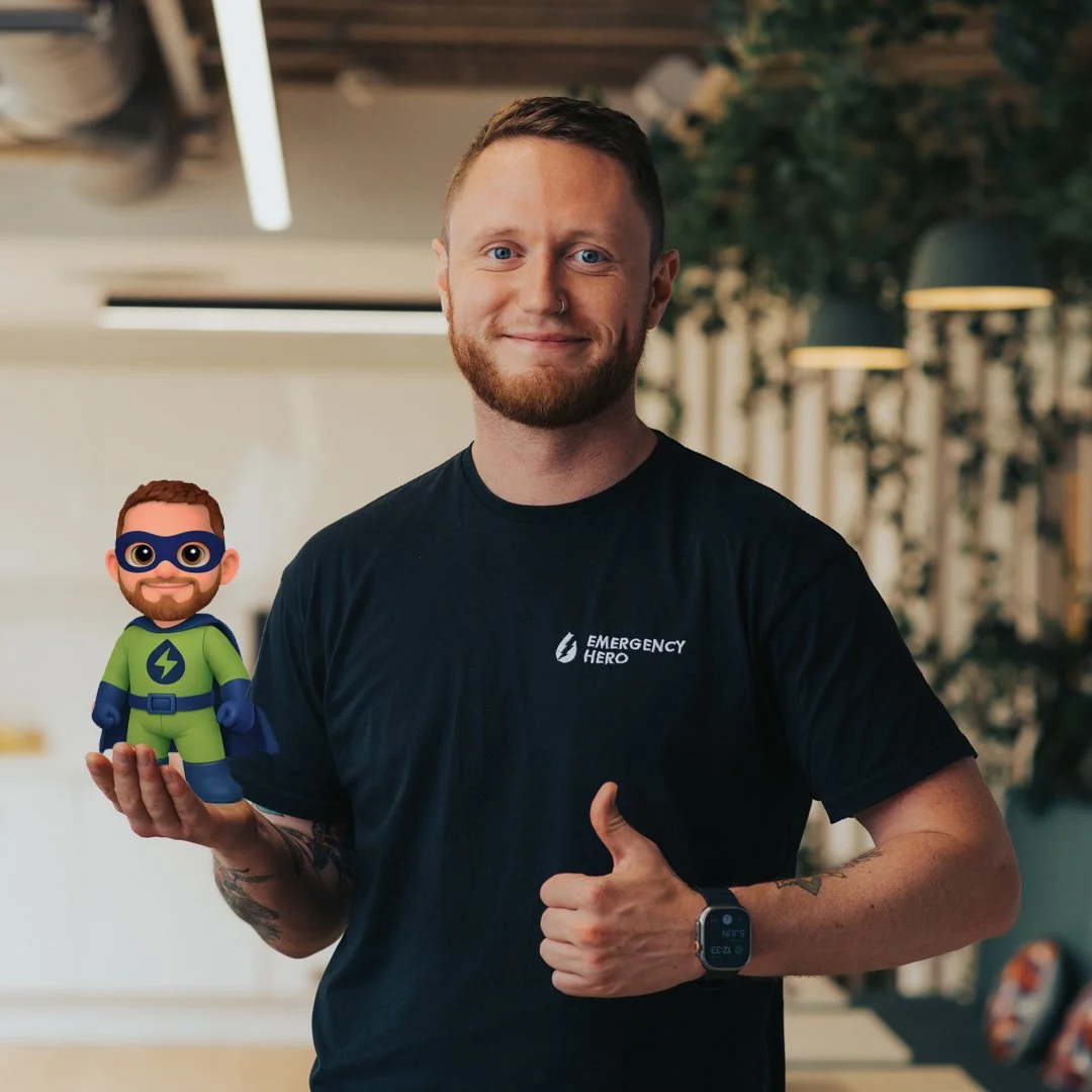 A smiling man with a beard and tattoos on his arm is giving a thumbs-up and holding a small superhero figurine. He is wearing a black T-shirt with the logo 'Emergency Hero' and is in an indoor setting with greenery in the background.