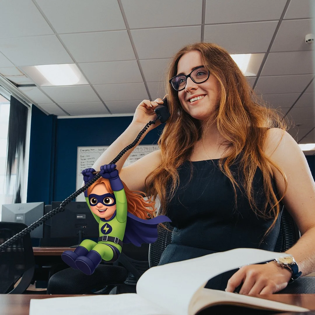 A woman with long red hair and glasses smiling while talking on a landline phone in an office setting. She is wearing a black sleeveless top and a wristwatch. There is a cartoon superhero character with glasses and a green costume holding a toolbox digitally edited into the image, positioned near the woman's arm.