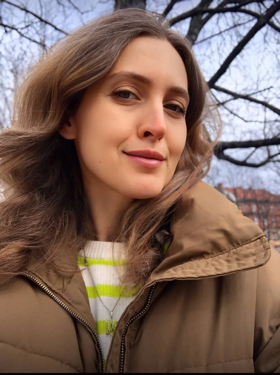 Frau im braunen Mantel im Freien, kahlköpfige Bäume im Hintergrund.