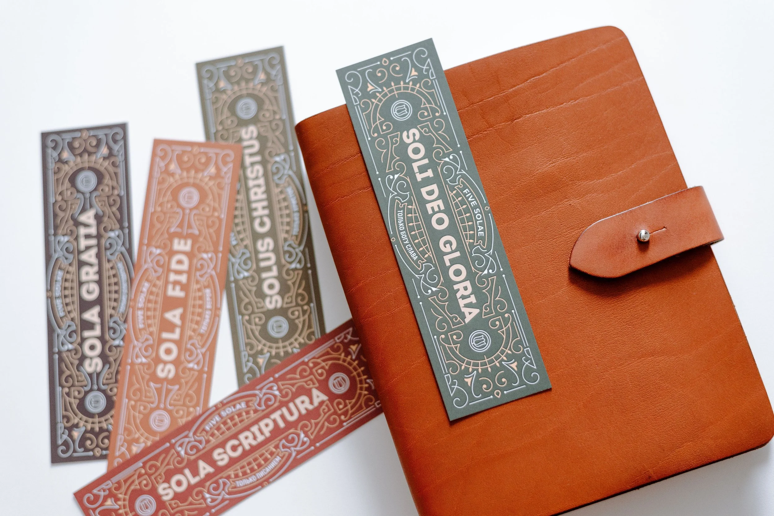 Eine braune Ledermappe mit fünf Lesezeichen in verschiedenen Farben und stilisierten Designs, die Sätze wie 'SOLA GRATIA', 'SOLA FIDE', 'SOLUS CHRISTUS', 'SOLI DEO GLORIA', und 'SOLA SCRIPTURA' tragen. Die Lesezeichen liegen teils auf der Mappe und teils daneben auf einem weißen Untergrund.