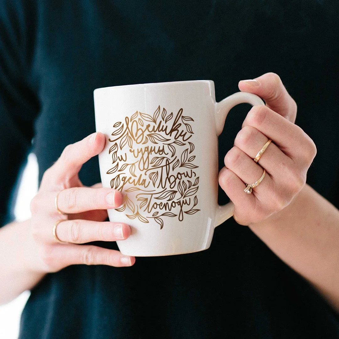Person hält eine weiße Tasse mit goldener Kalligrafie und Blattverzierungen. Die Hände tragen Ringe.