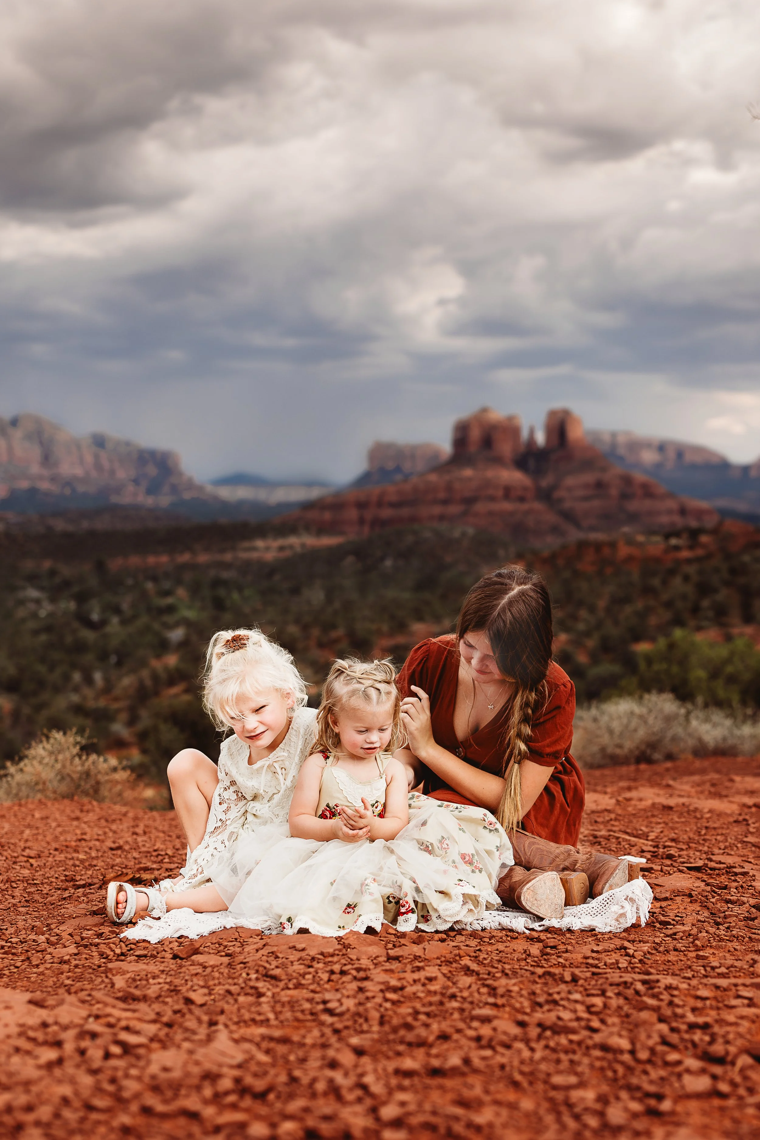 A woman and three young children sitting on a blanket in a desert landscape with red rocks and mountains in the background, under cloudy skies. Family photography, Sedona arizona.