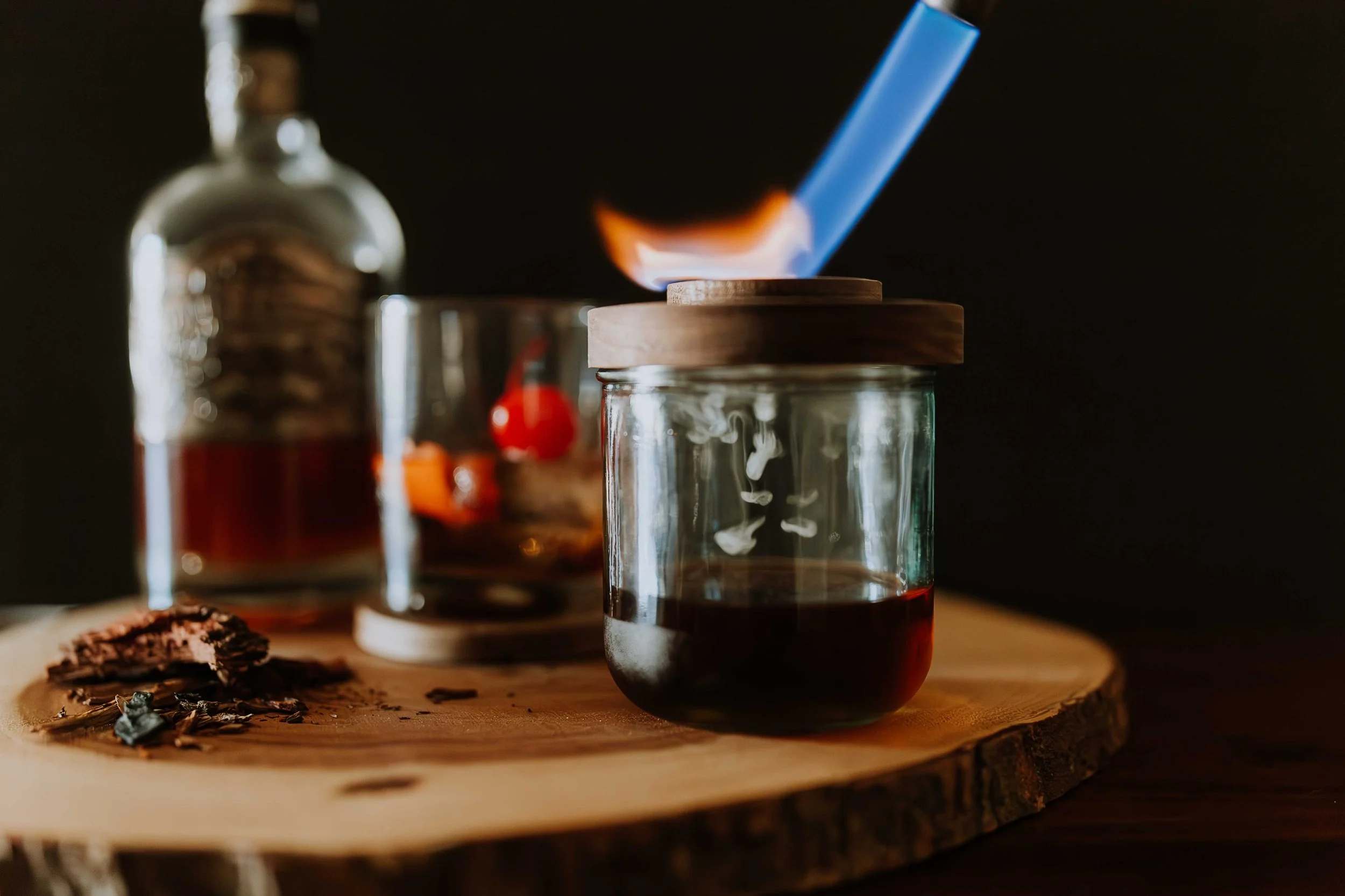 A small glass jar with a wooden lid contains a dark red substance, possibly jam or preserves, placed on a wooden surface. Behind, there are other jars, one with a partially visible red label and another smaller jar, along with some scattered dried tea leaves or herbs in the foreground. A blue glass straw is inserted into the jar with the wooden lid against a dark background.