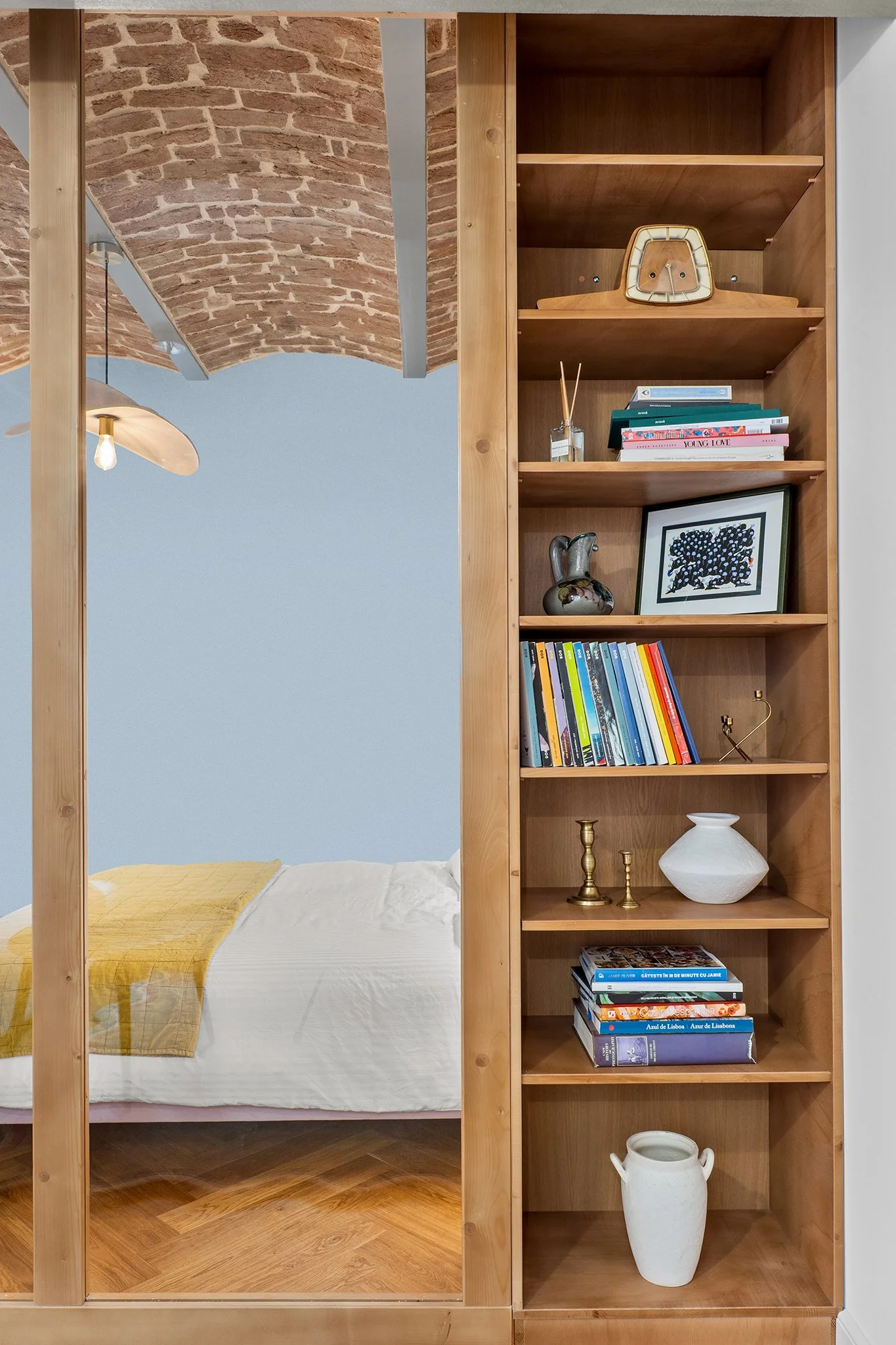 A view of a bedroom with a bed and a wooden bookshelf filled with books and decorative items, visible through a mirror.