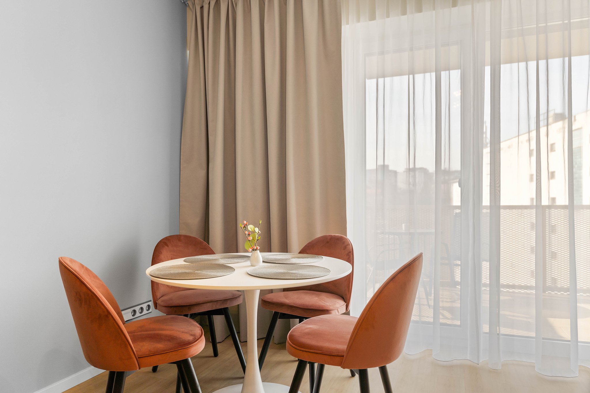 Modern dining area with a round white table, four orange chairs, and a small vase with flowers. Beige curtains and sheer drapes cover large windows.