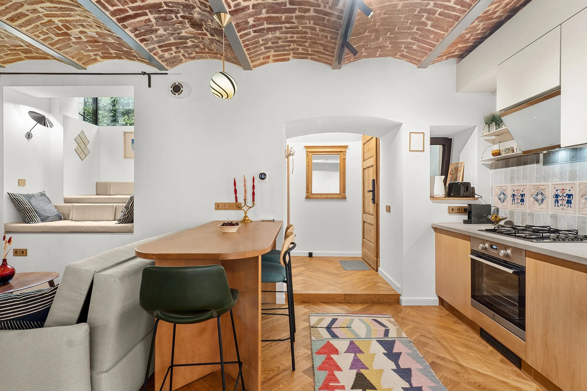 Interior view of a modern kitchen with wooden cabinets, a gas stove, and colorful decorative tiles, overlooking a cozy living area with a built-in window seat and a brick arched ceiling.