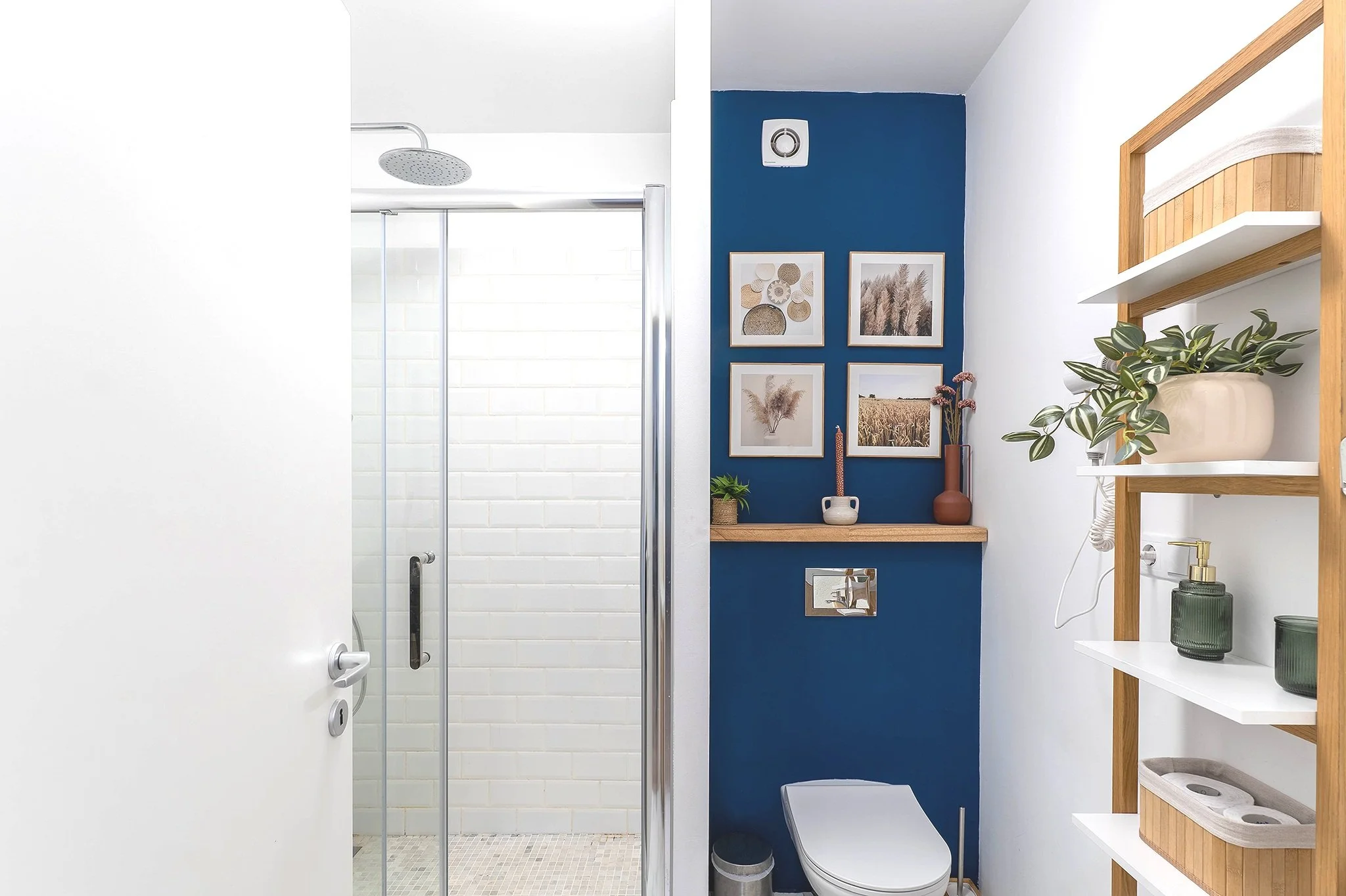 Modern bathroom with blue accent wall, framed botanical prints, white shelves, potted plant, toilet, and walk-in shower with glass door.