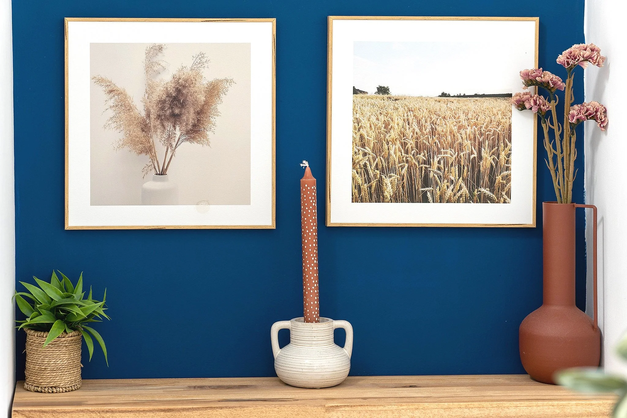 Interior decor with two framed pictures, a tall candle in a ceramic holder, a small potted plant, and pink flowers in a tall vase on a wooden surface against a blue wall.